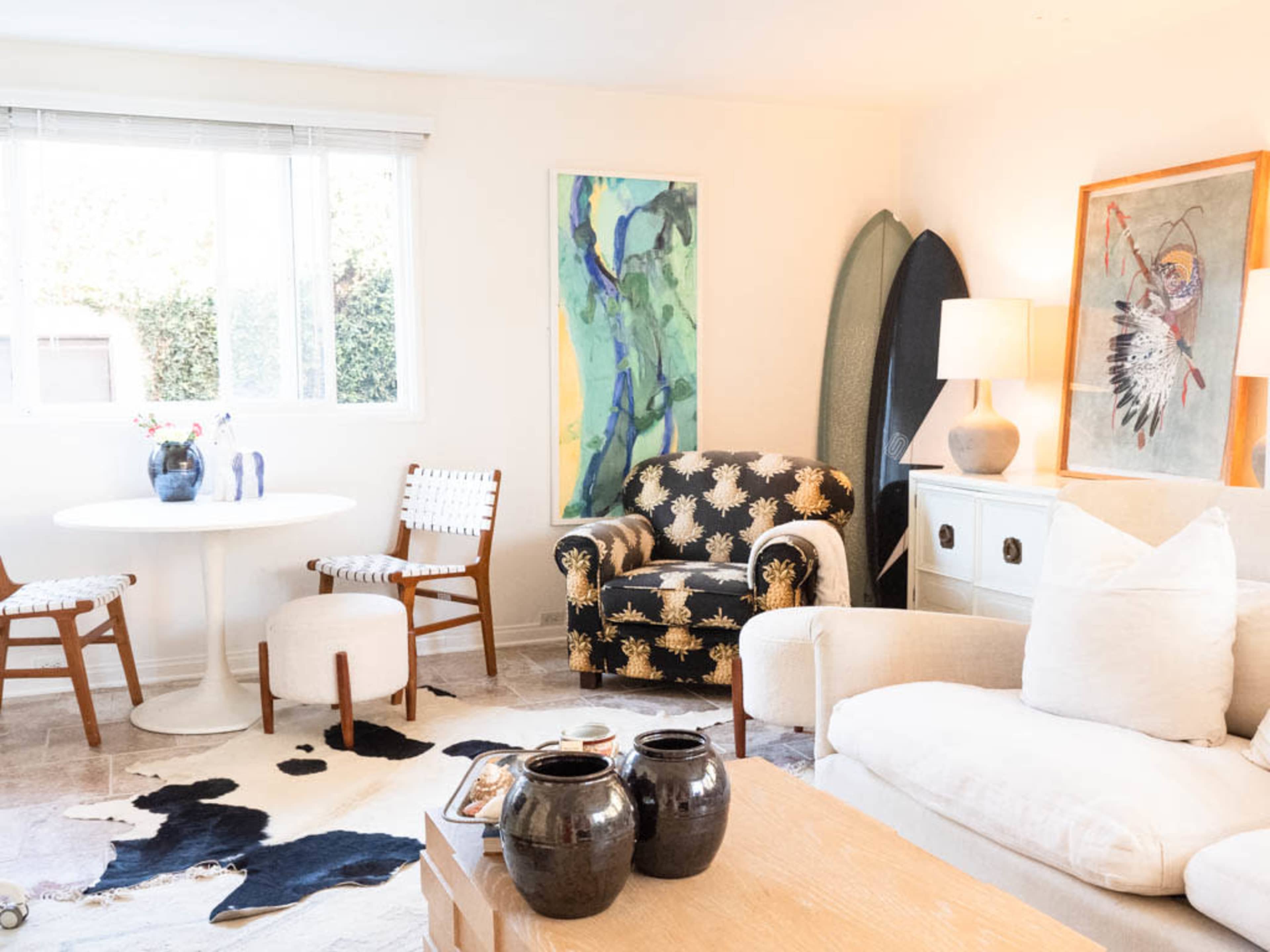 The image shows a bright living room featuring a round dining table, a patterned armchair, a light-colored sofa, a wooden coffee table with decorative vases, and artwork on the walls.