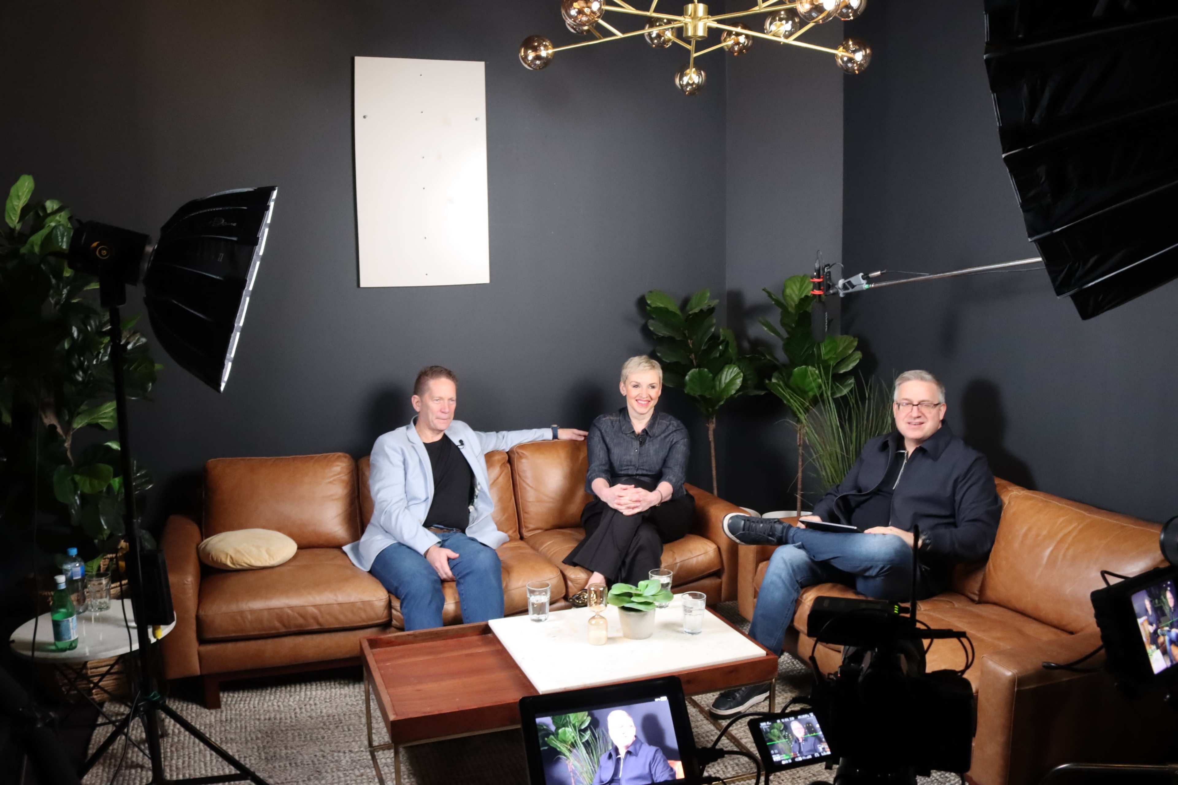 Three people are seated on a brown leather couch in a studio setting, surrounded by plants and cameras, with bright lighting overhead.