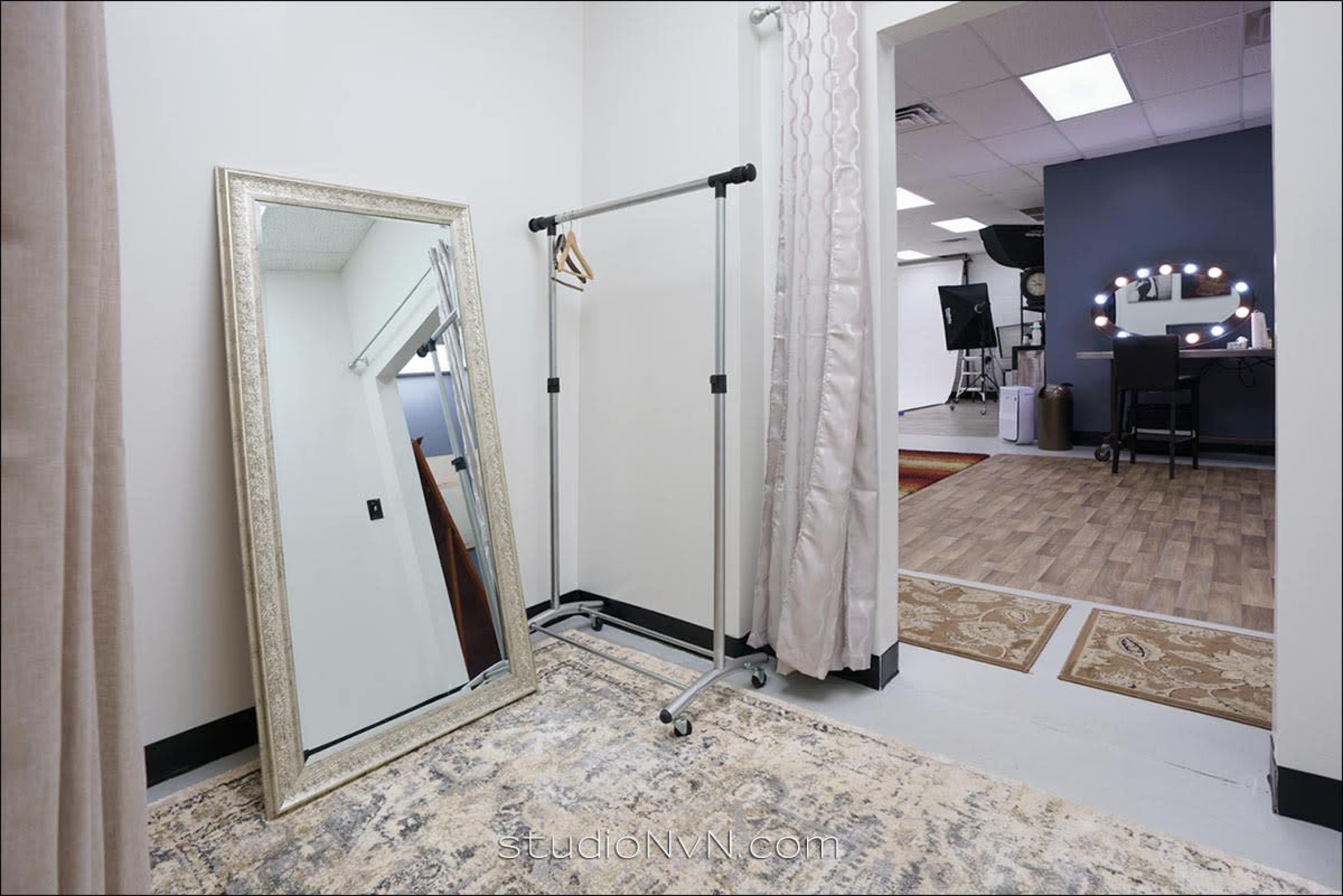 The image shows a bright interior space featuring a large mirror, a clothing rack, and a gray wall with illuminated vanity mirrors in the background.