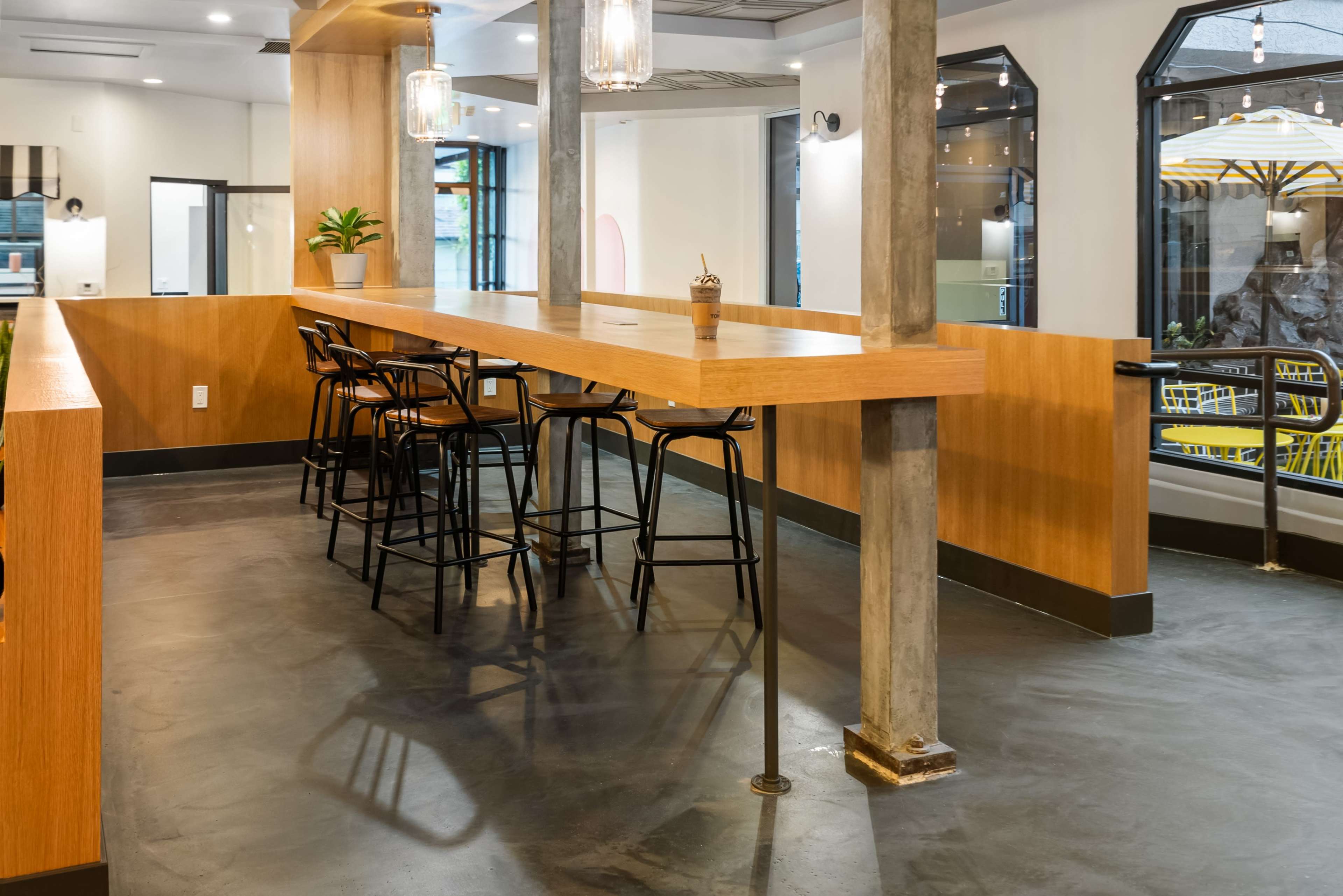 A modern indoor café space featuring wooden counters, metal bar stools, and large windows.