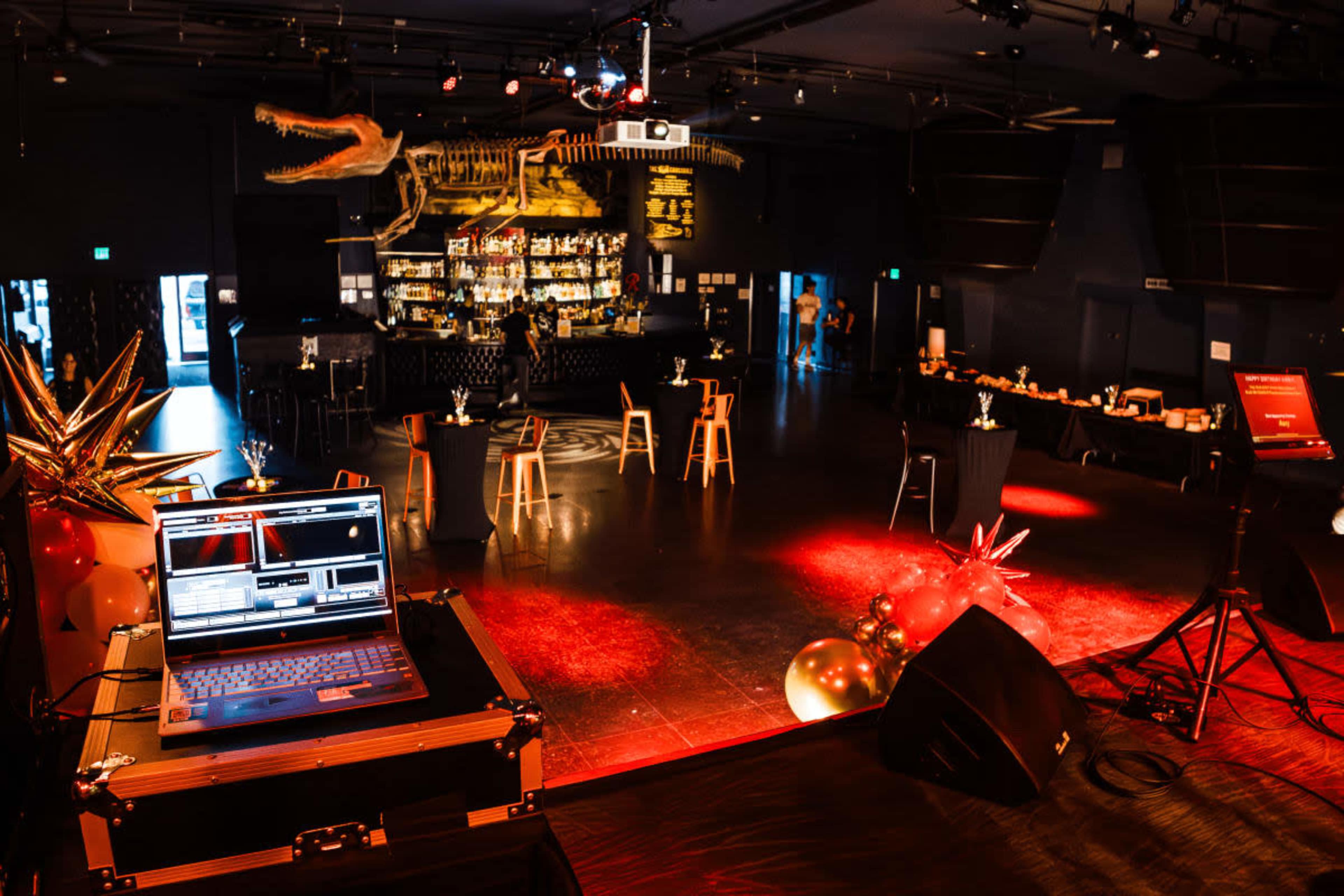 The image shows a dimly lit event venue featuring a bar, seating arrangements, a DJ setup, and decorative elements like balloons and stars.