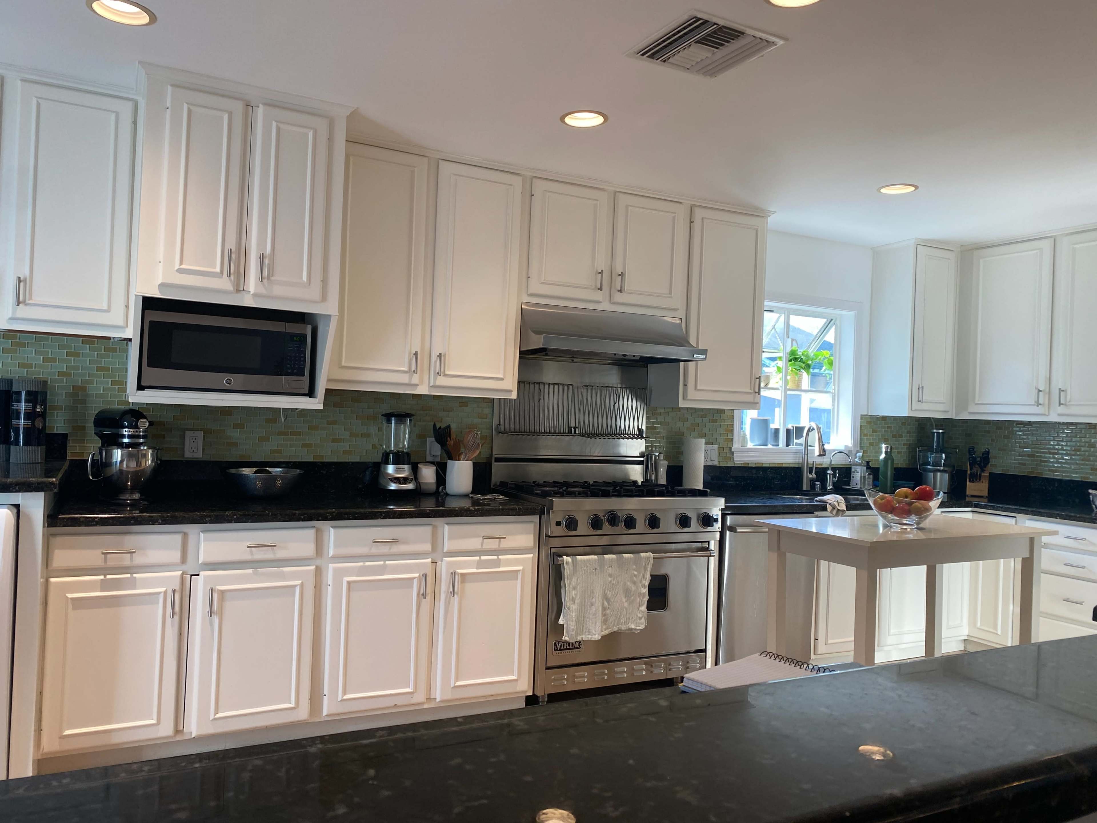 The image shows a modern kitchen featuring white cabinets, black countertops, stainless steel appliances, and a small island with a bowl of fruit.
