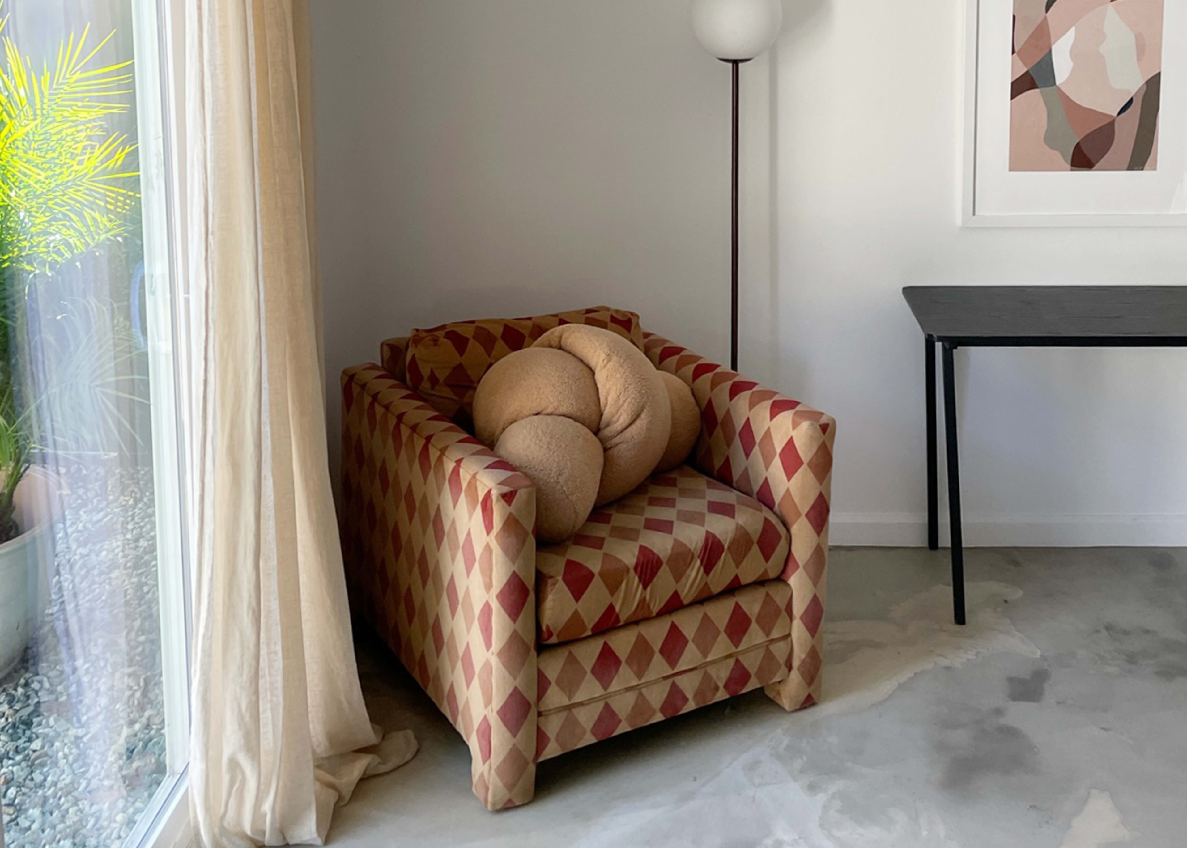 A patterned armchair with a large, round cushion sits in a corner next to a lamp and a table in a minimalist room.