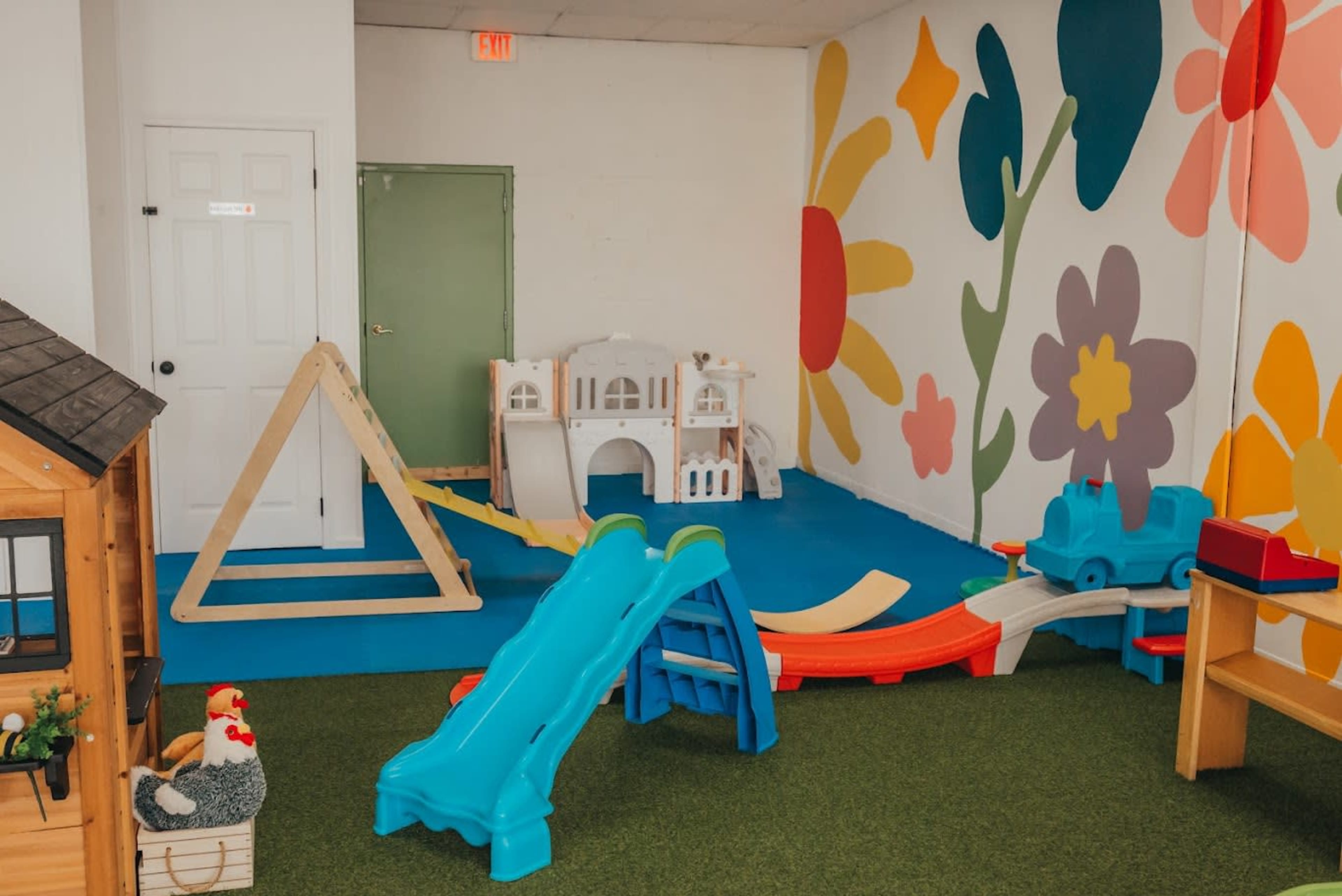 The image shows a colorful playroom featuring a slide, climbing structure, toy vehicles, and decorative flower murals on the walls.