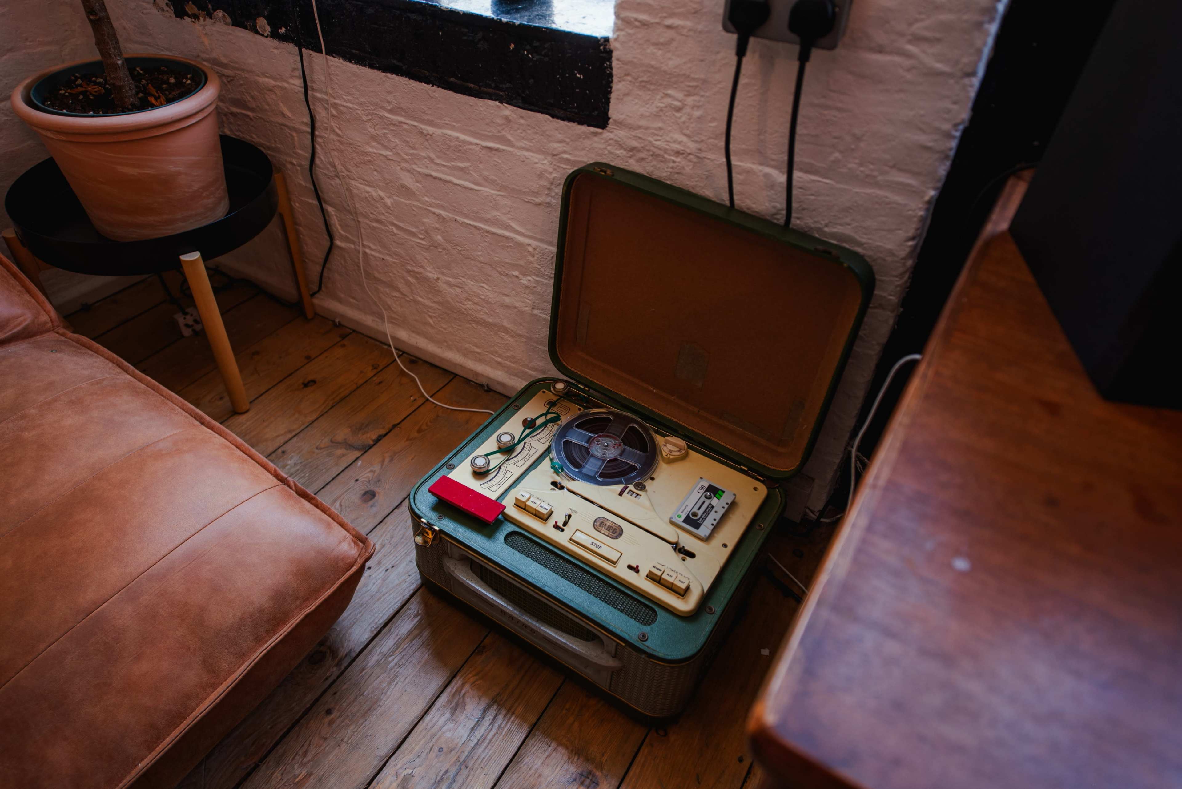 A vintage reel-to-reel tape recorder sits on the wooden floor next to a leather couch and a potted plant.