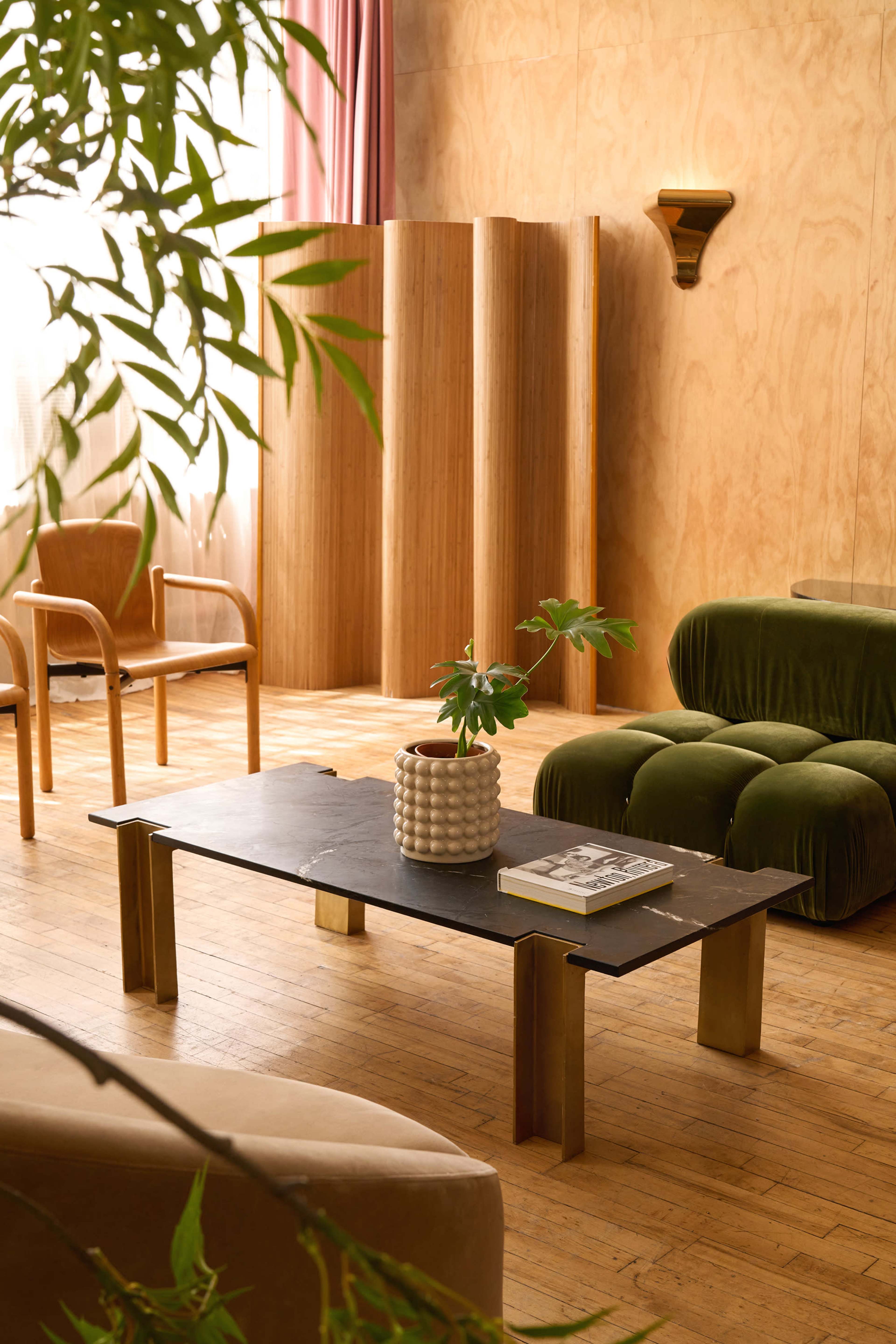The image depicts a modern living space featuring a wooden floor, a green velvet sofa, a light-colored armchair, a coffee table with a black surface, and a potted plant on the table.