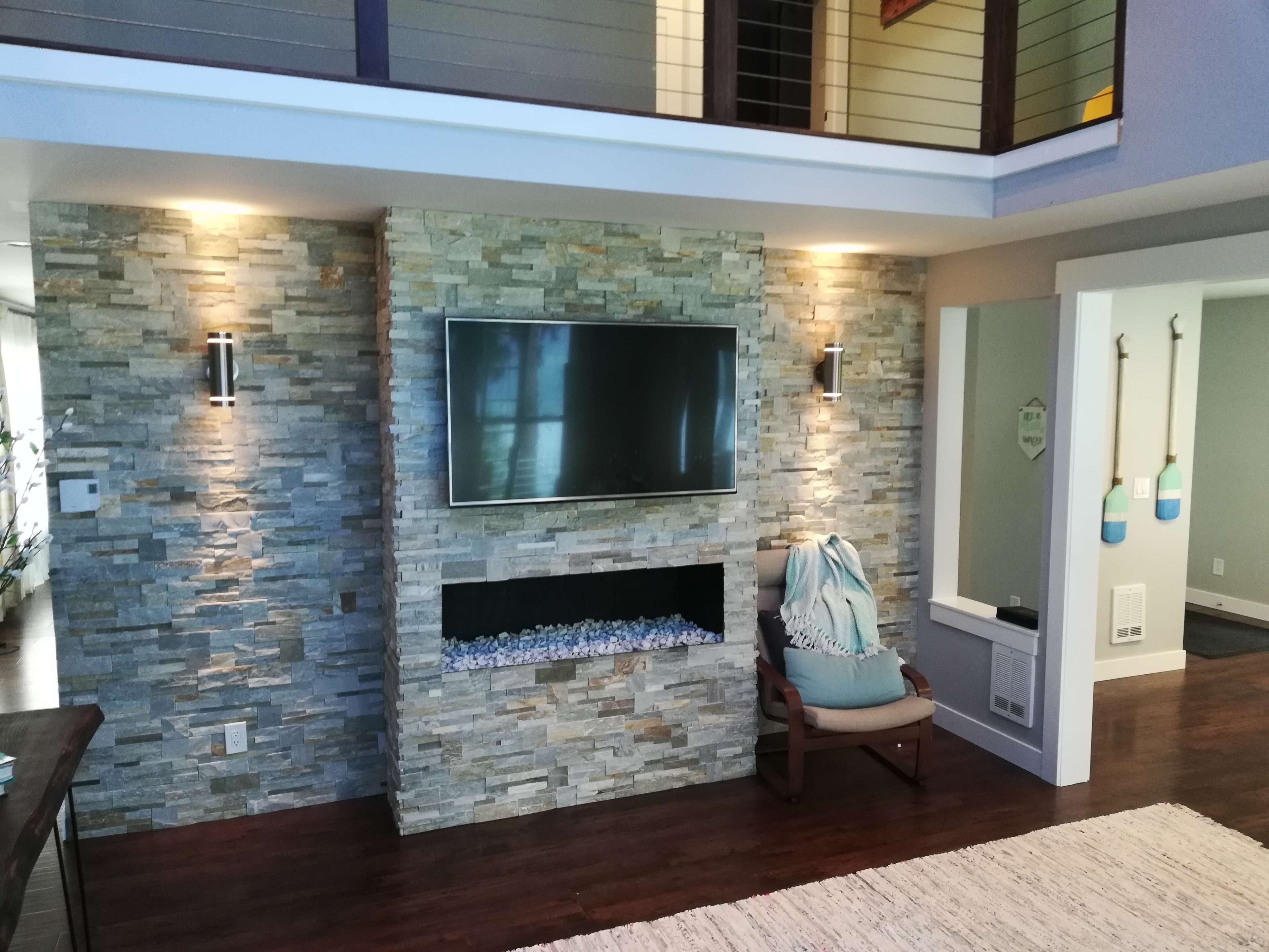 The scene features a modern living room with a stone accent wall, a mounted television, and a cozy reading chair beside a fireplace-like structure.