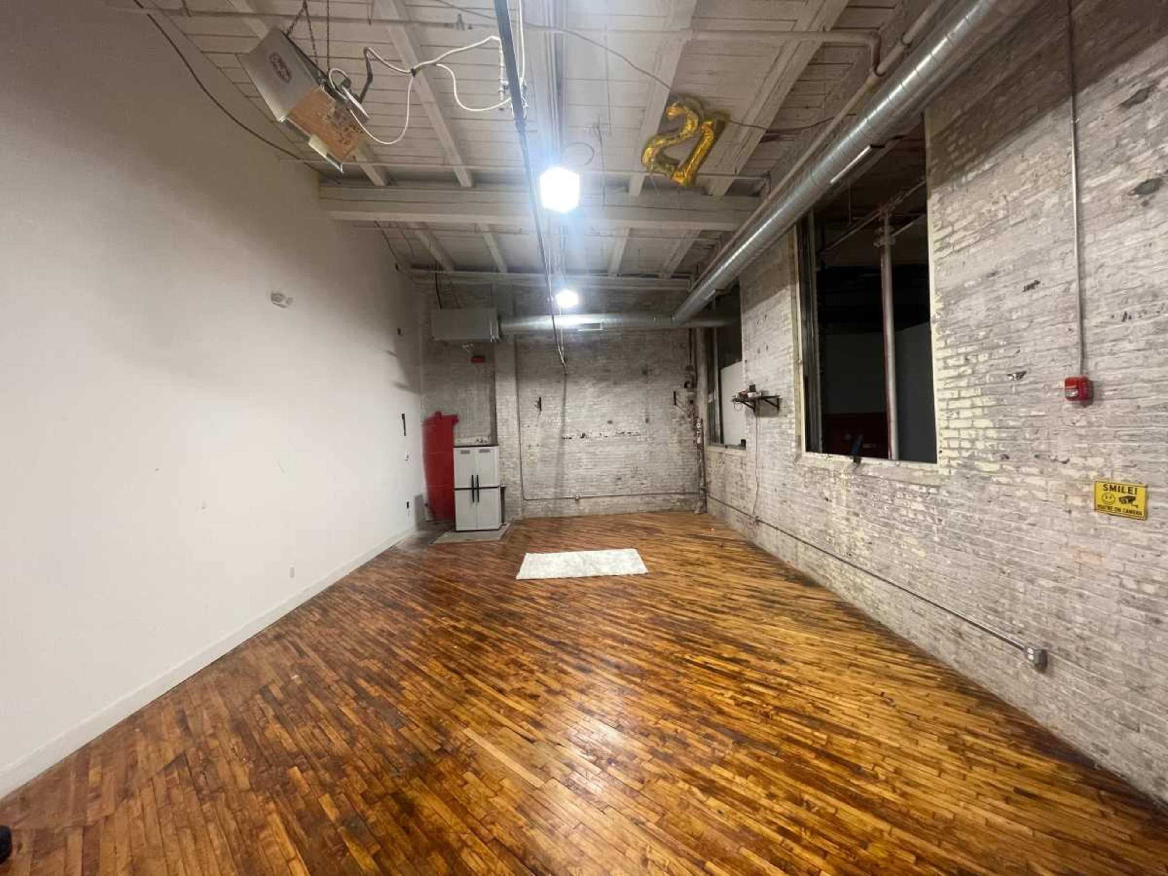 The image shows a spacious, undecorated room with hardwood floors, exposed brick walls, overhead lighting, and minimal furnishings.