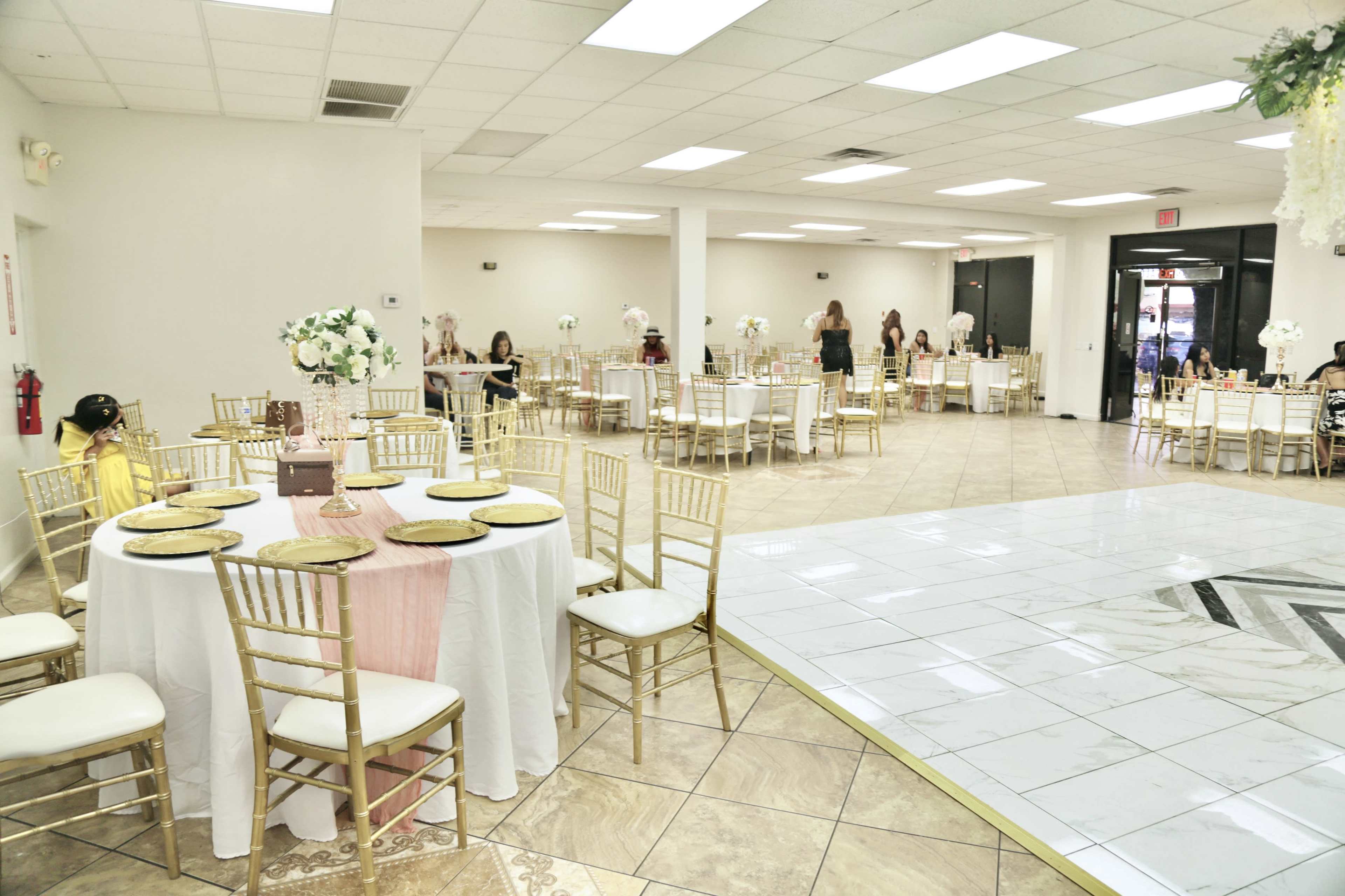 The image shows an elegantly set event space with round tables covered in white tablecloths and gold chairs, featuring floral centerpieces and a large open floor area.