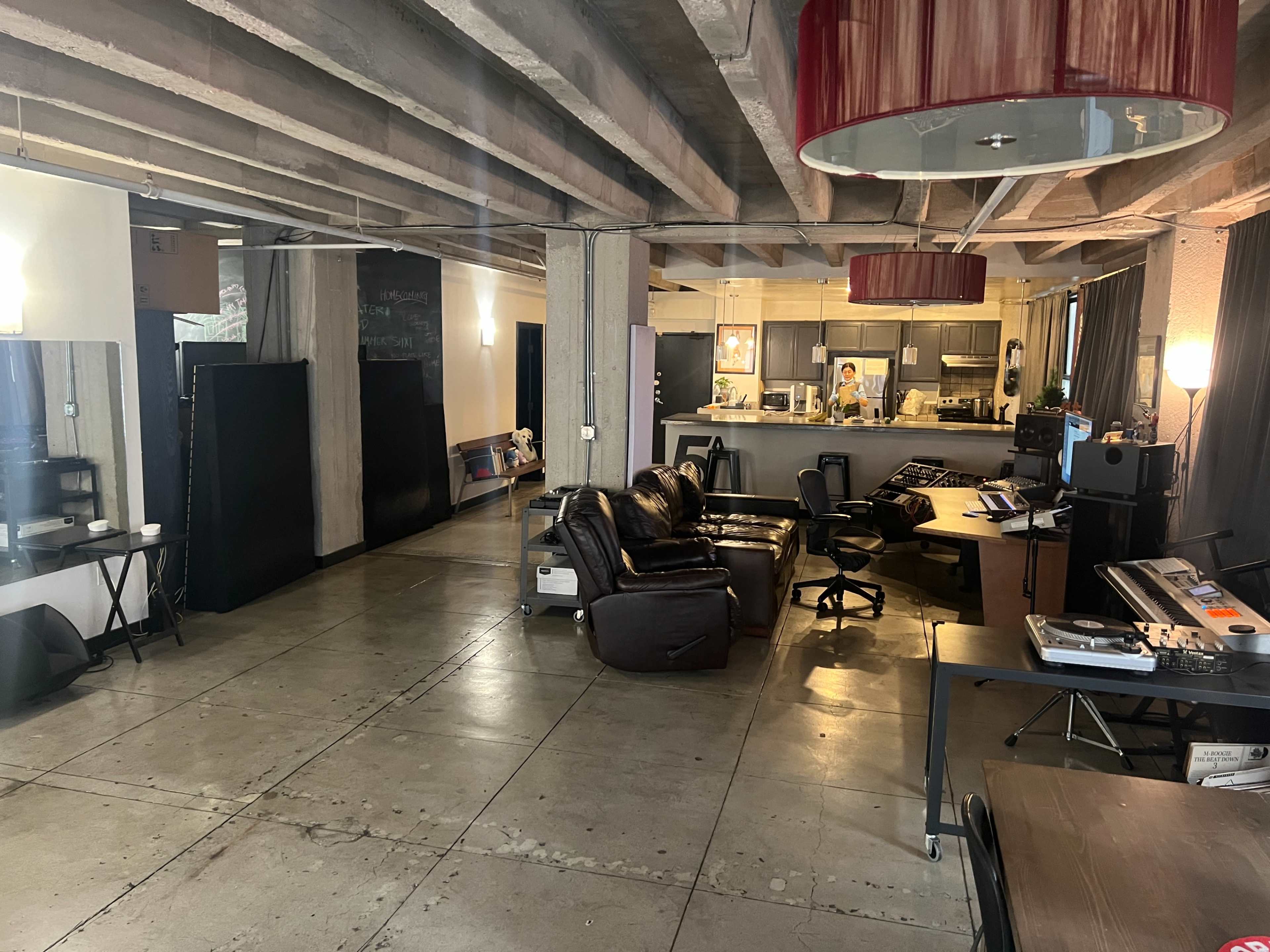 The image shows a spacious open-plan living area featuring a kitchen, seating area with leather couches, and various workspaces with modern furniture.