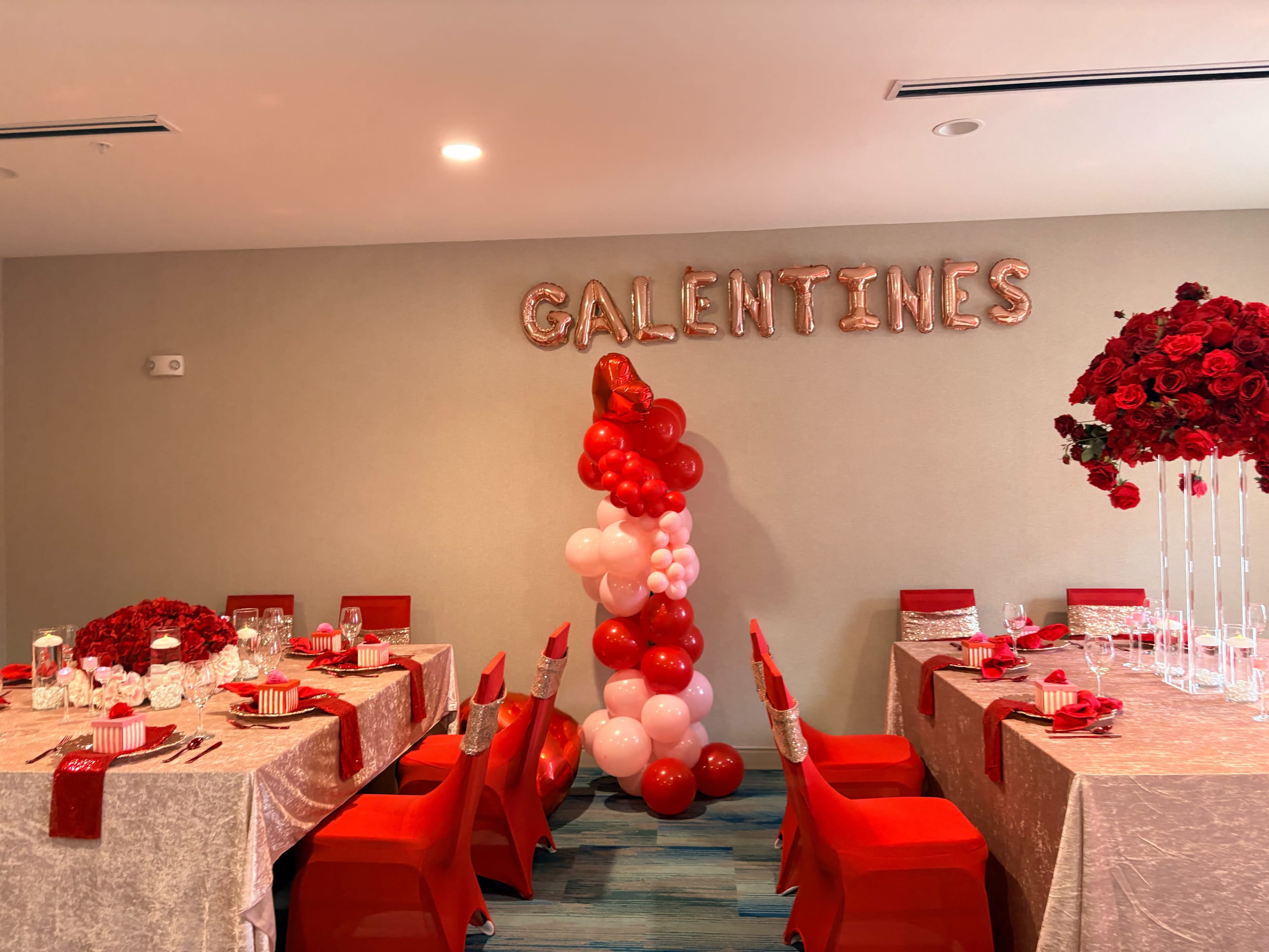 The image shows a decorated event space for a Galentine's celebration, featuring tables set with red and pink accents, balloons, and floral centerpieces.