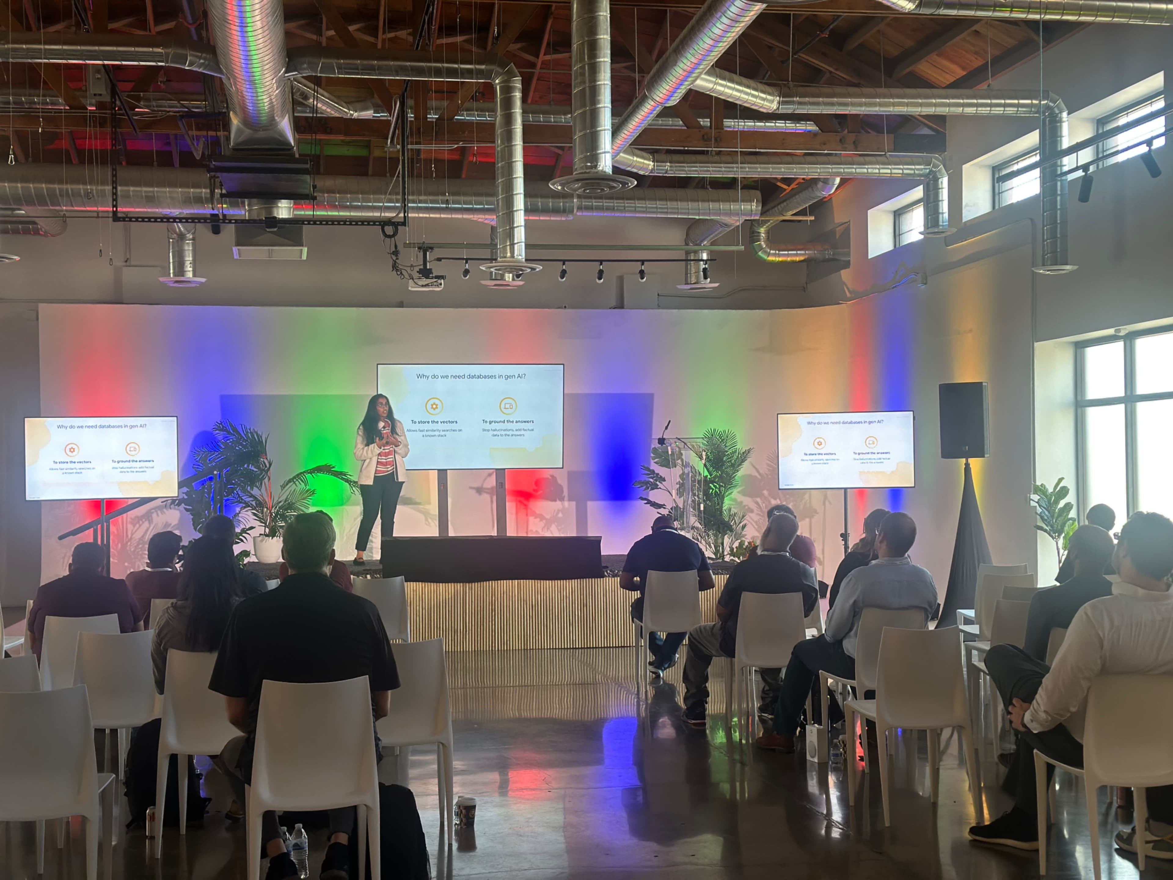 A speaker presents information to an audience in a brightly lit venue with colorful lighting and multiple screens.