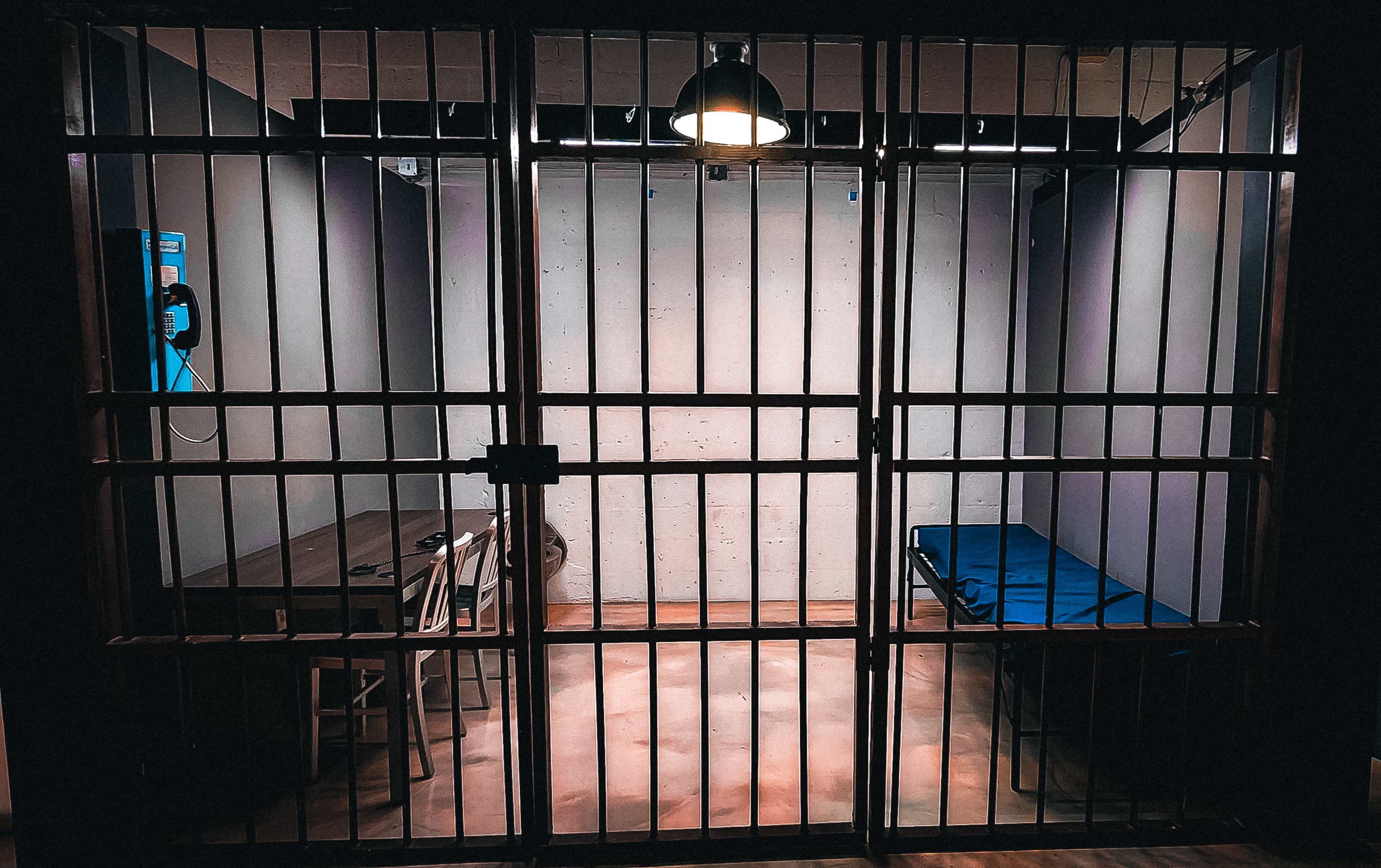 The image shows a dimly lit jail cell with metal bars, containing a bed, a table with chairs, and a telephone on the wall.