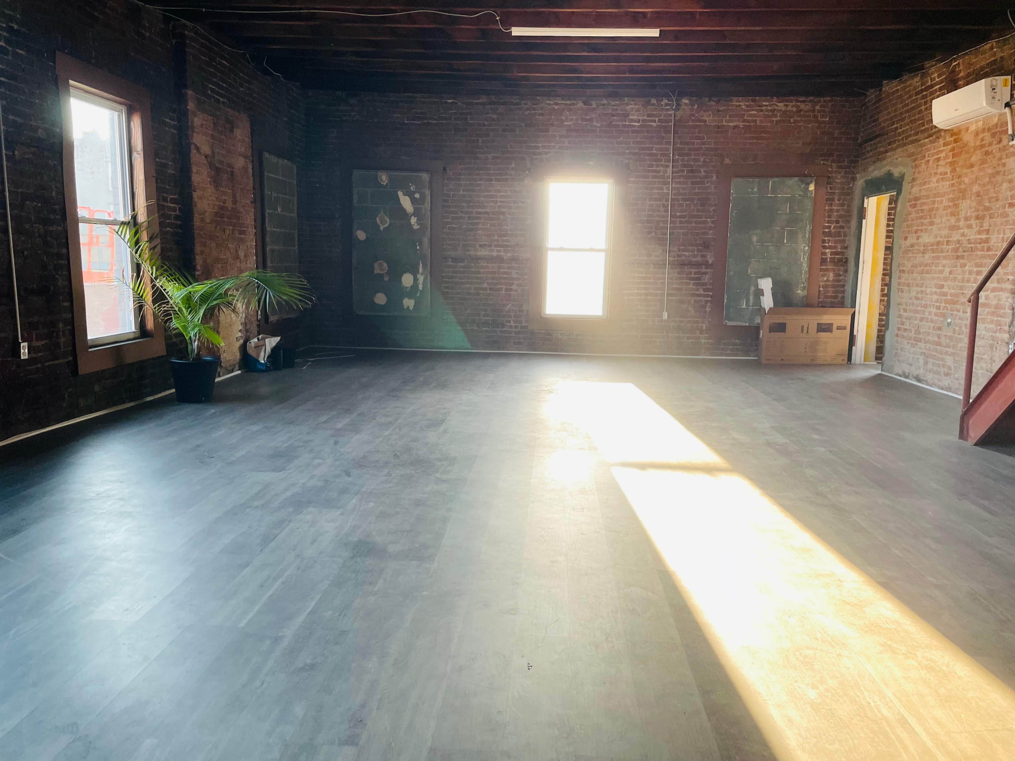 An empty room with exposed brick walls, large windows allowing sunlight to stream in, and a potted plant in one corner.