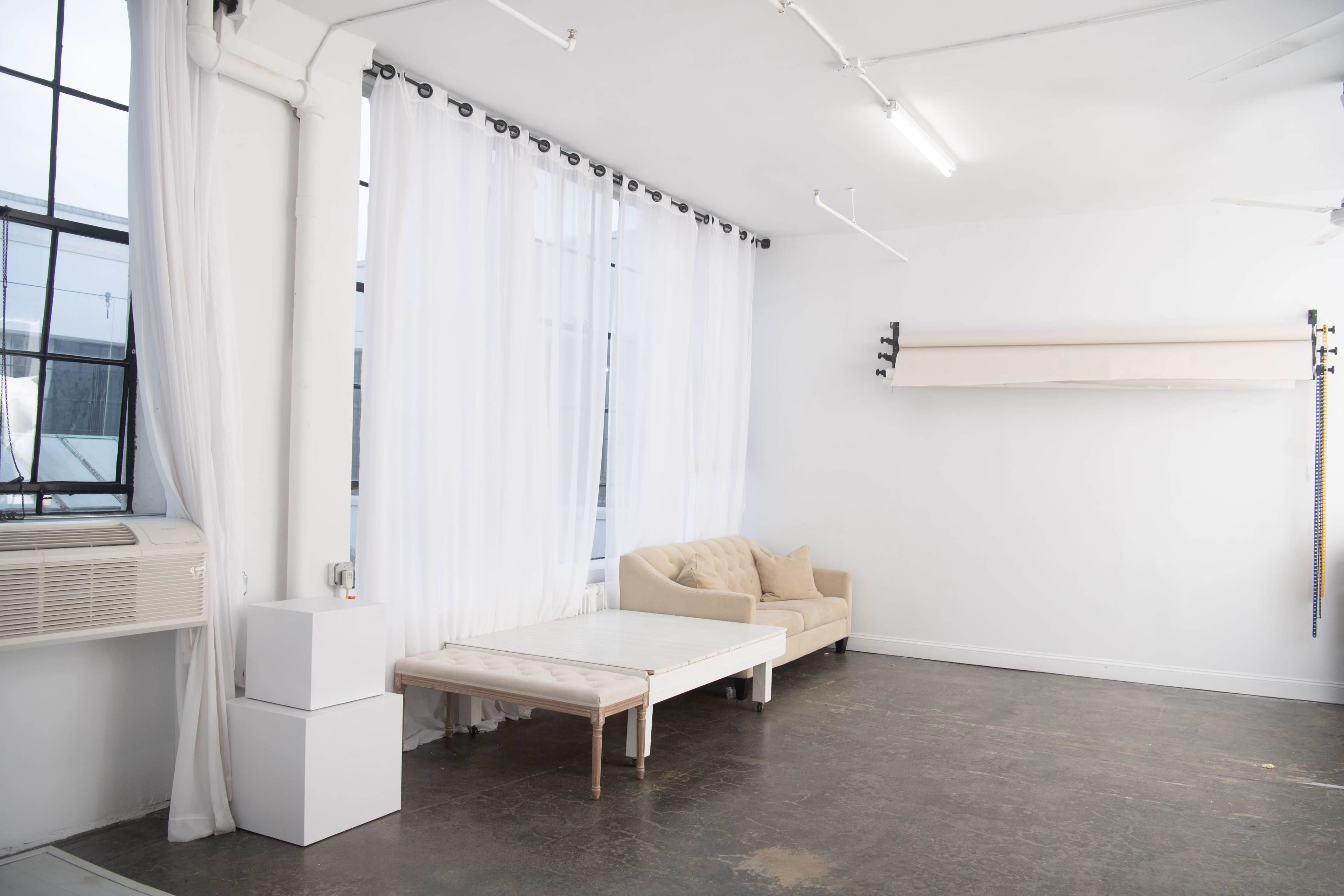 A bright, empty room features a light-colored couch, a small table, and large windows with sheer white curtains.