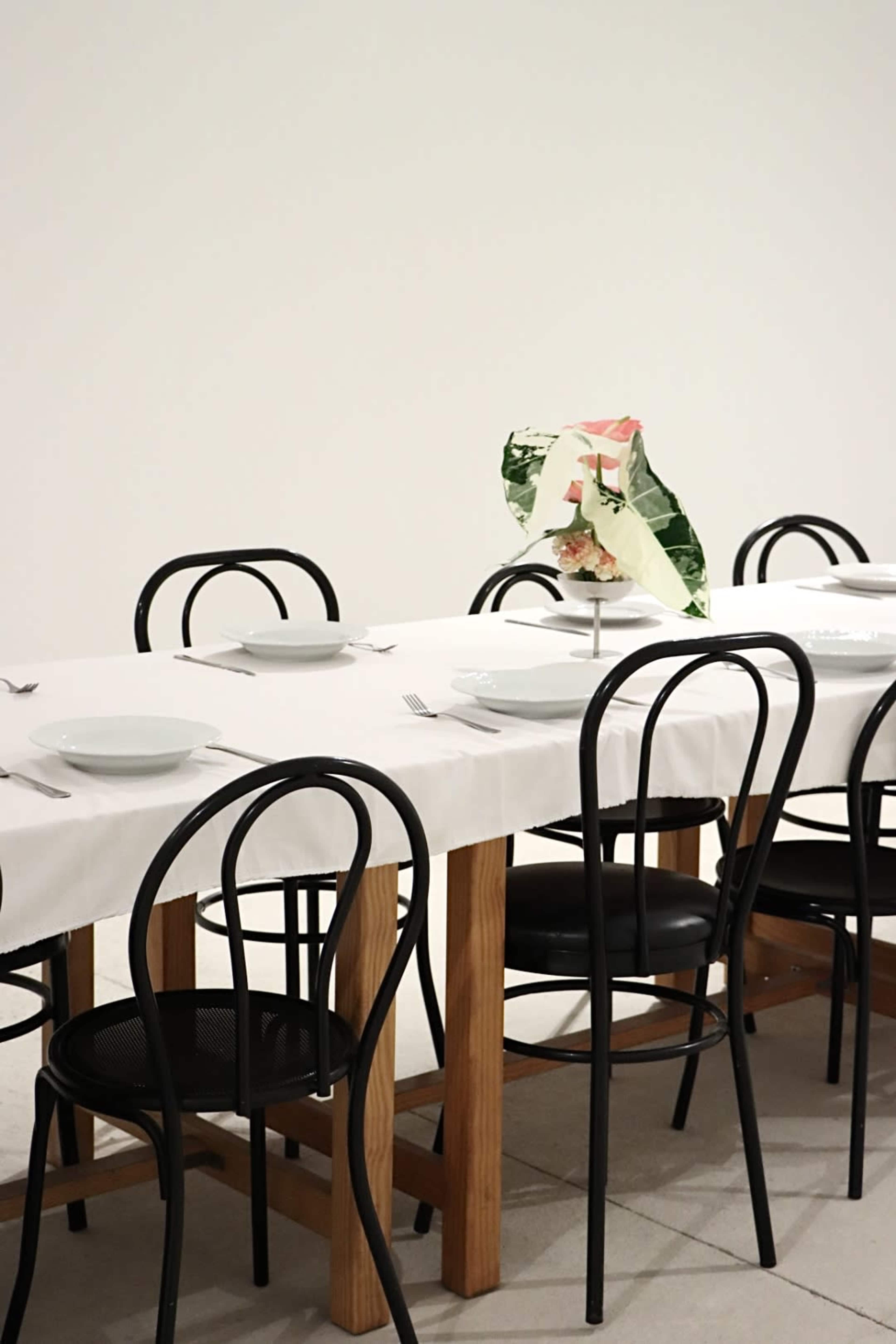 A long dining table with a white tablecloth is set with plates and cutlery, accompanied by a single decorative arrangement on top, surrounded by black chairs.