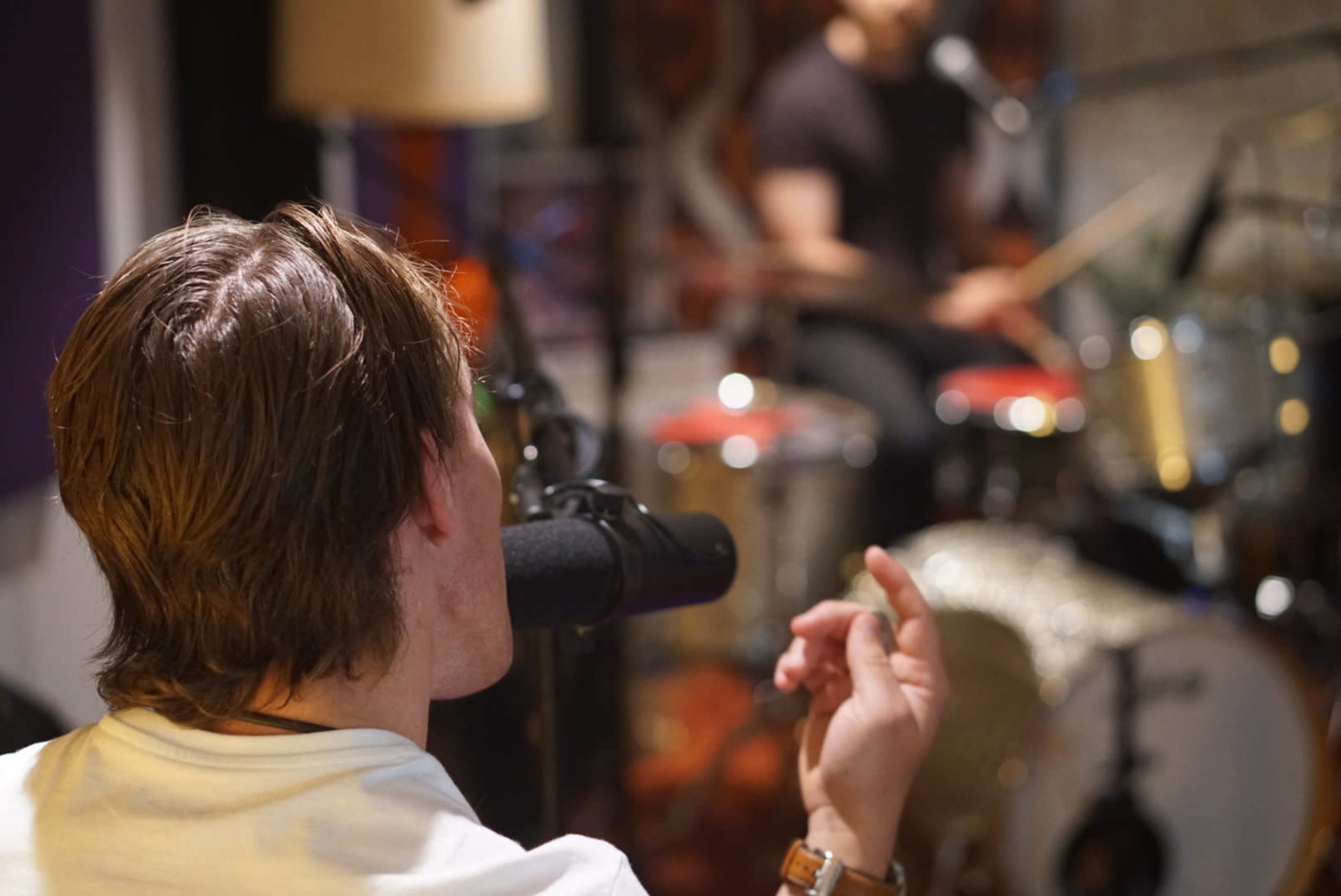 A person is seated in front of a microphone while a drummer is positioned in the background, playing a set of drums.