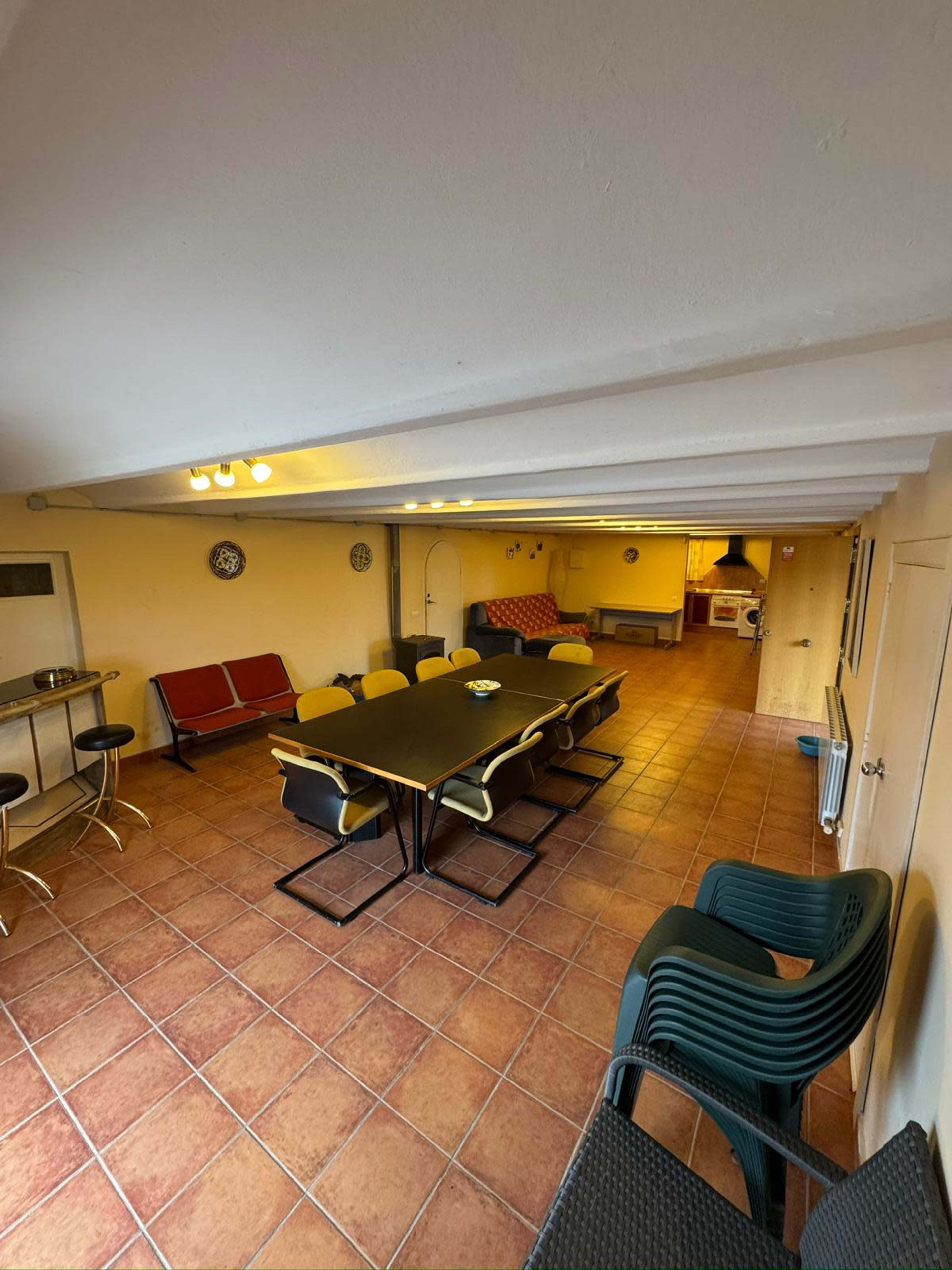 The image shows a spacious indoor room with a large table surrounded by chairs, a kitchenette area, and a sitting area with couches.