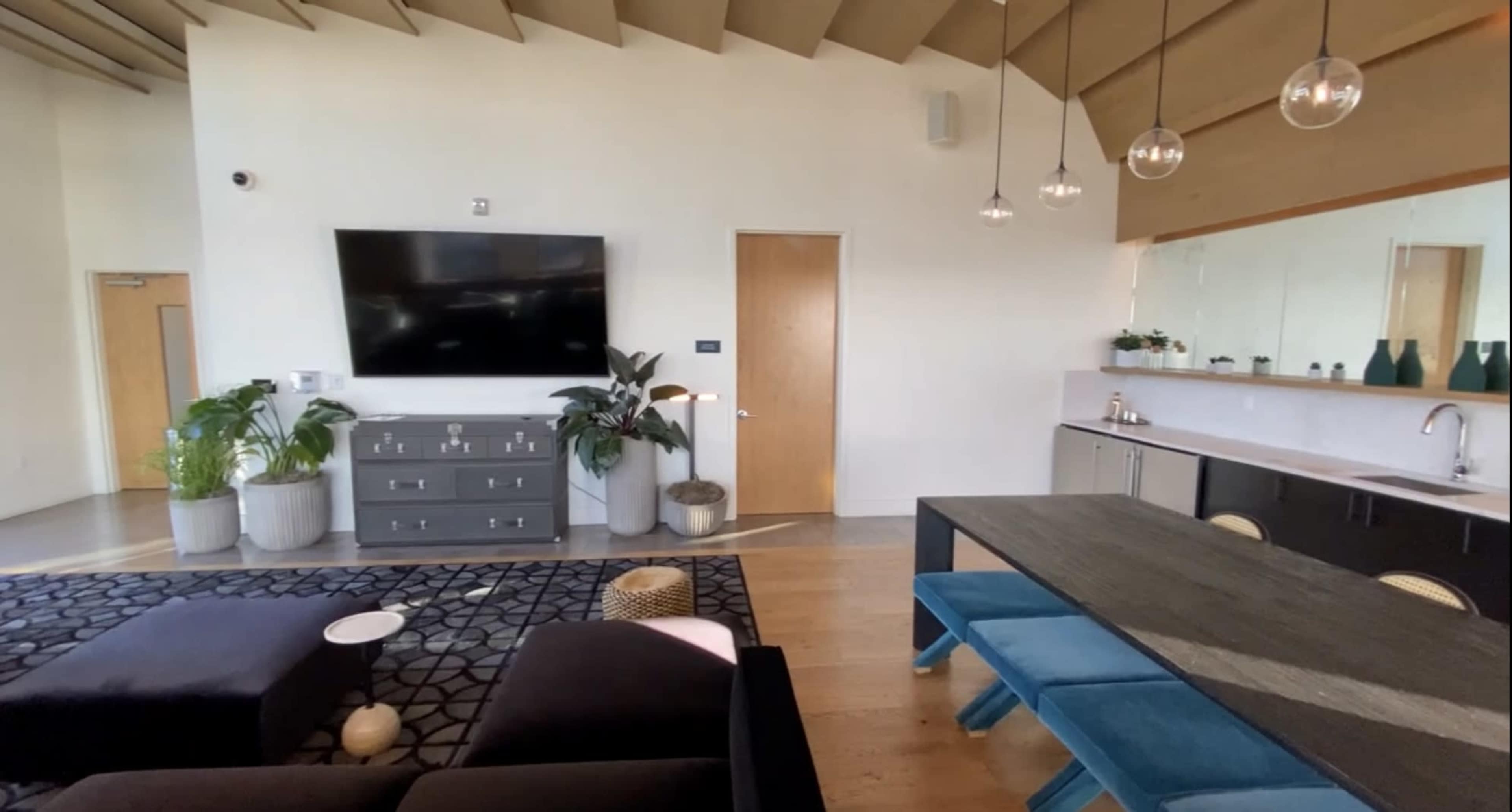 A modern living space featuring a dark sofa, a large television mounted on the wall, a dining table with blue chairs, and plants in decorative pots.