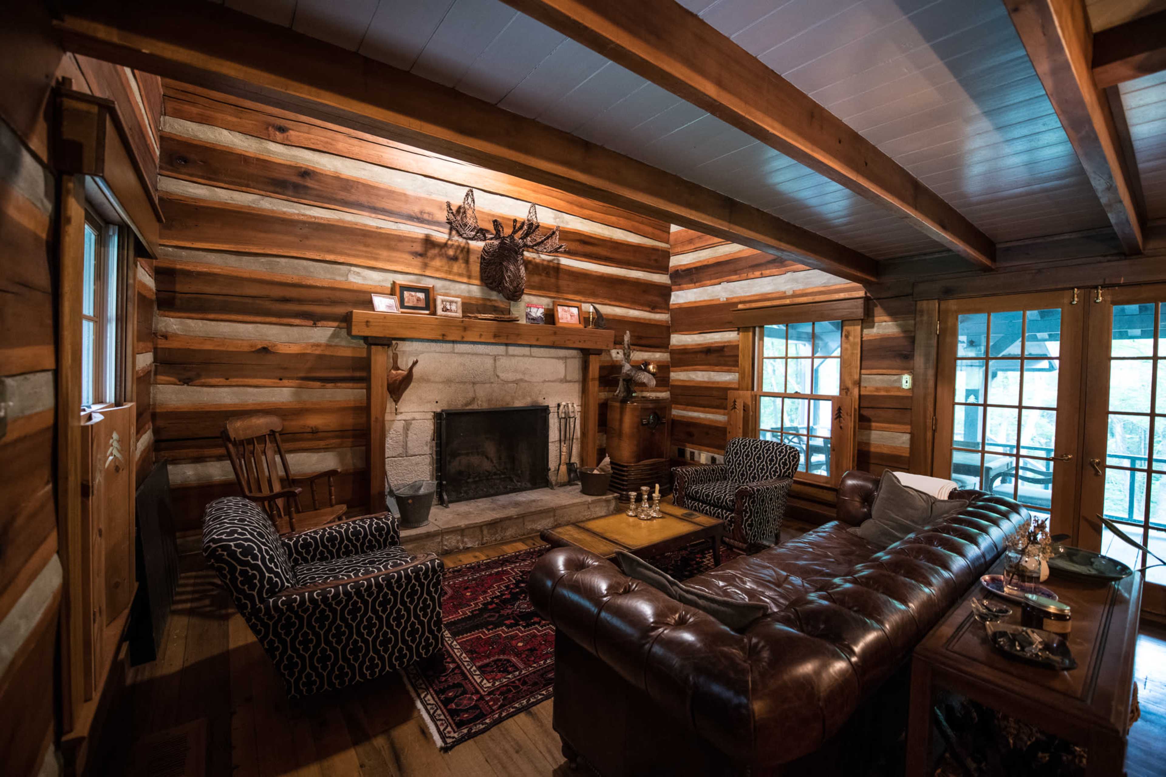 The image shows a log cabin interior featuring a leather sofa, a stone fireplace, and wooden beams on the ceiling.