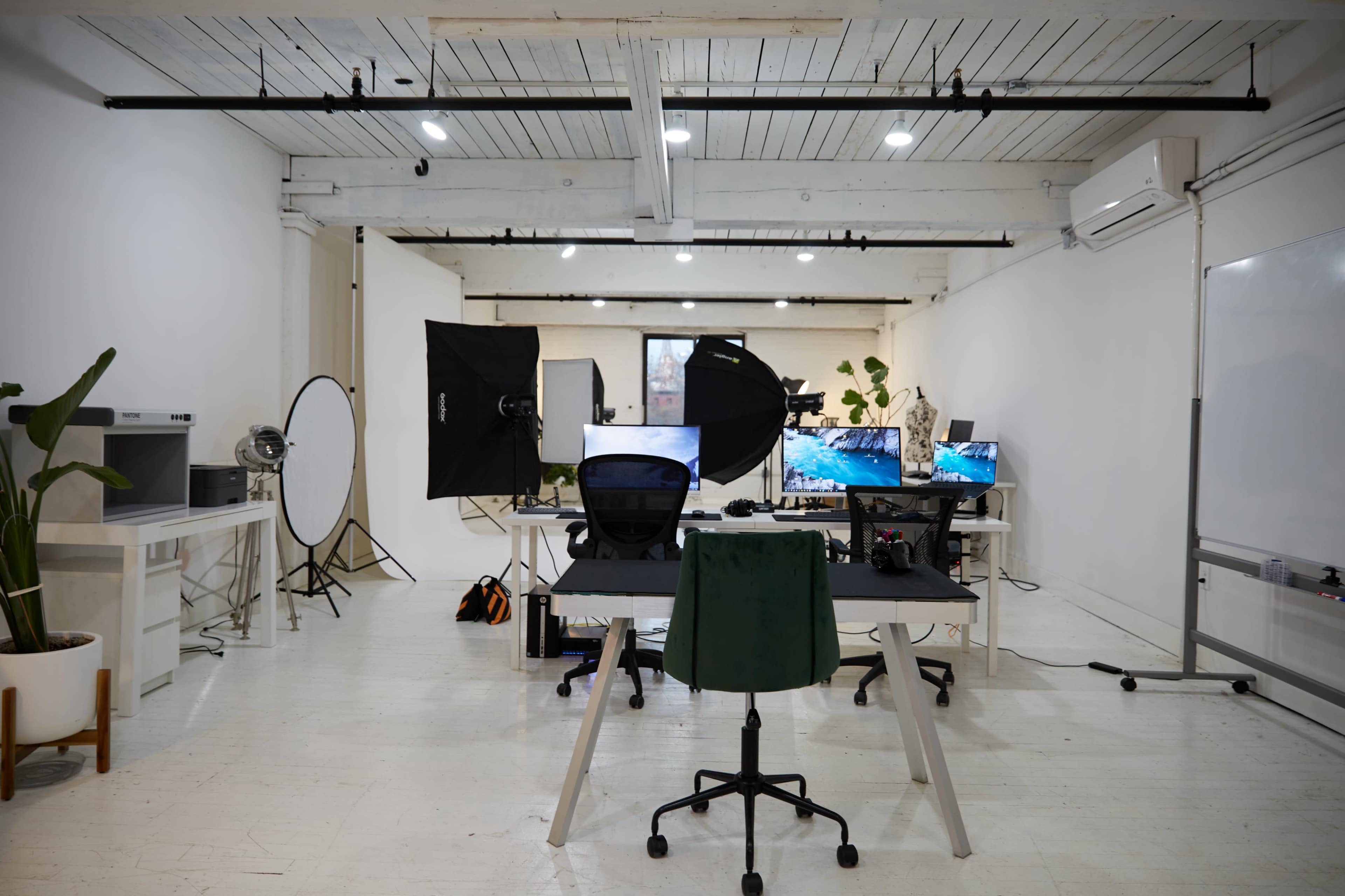 A well-lit studio workspace features multiple computer monitors, photography equipment, and a desk surrounded by chairs.