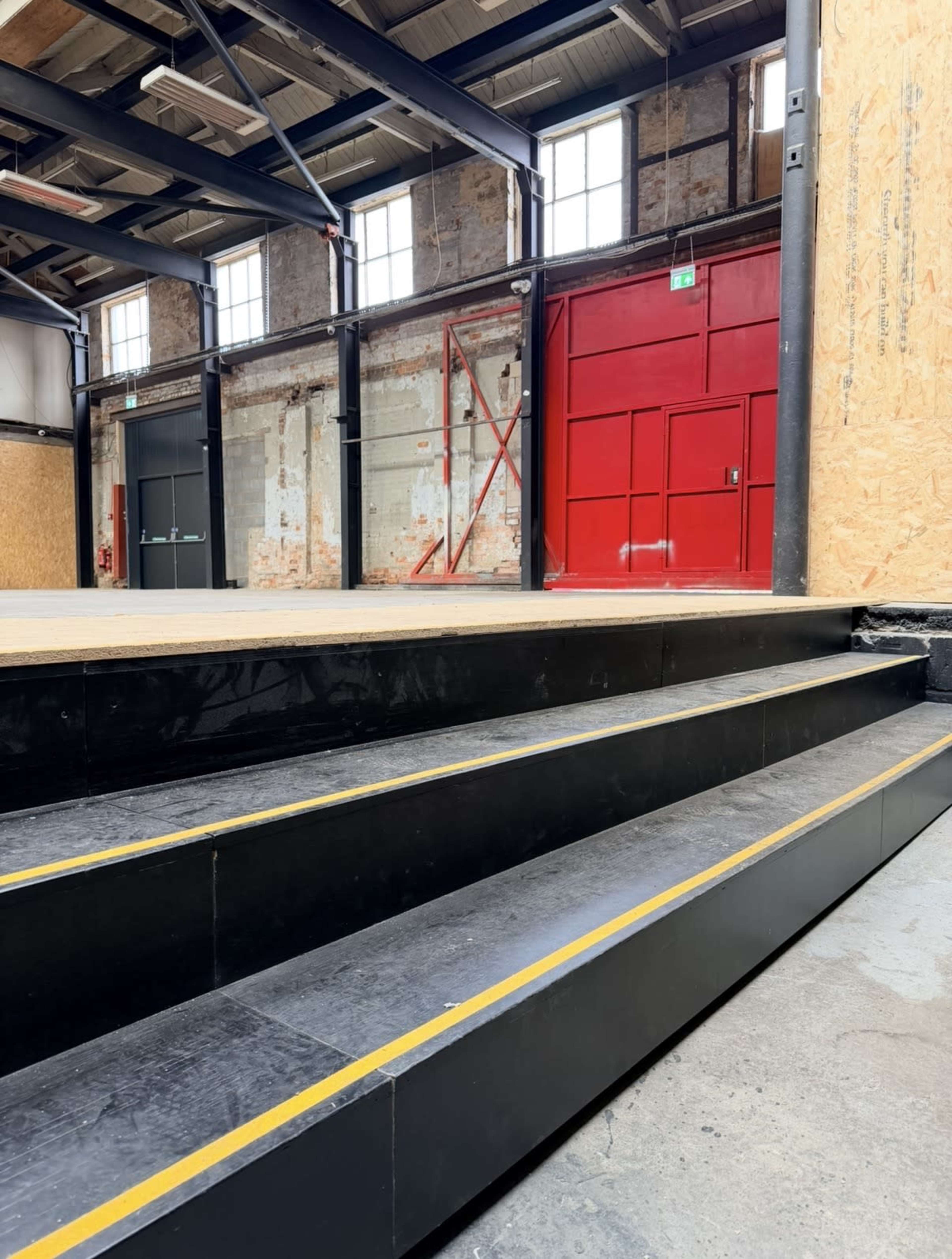The image shows a flight of black steps with yellow markings leading up to a wooden platform in an industrial-style building, featuring large windows and a red door in the background.