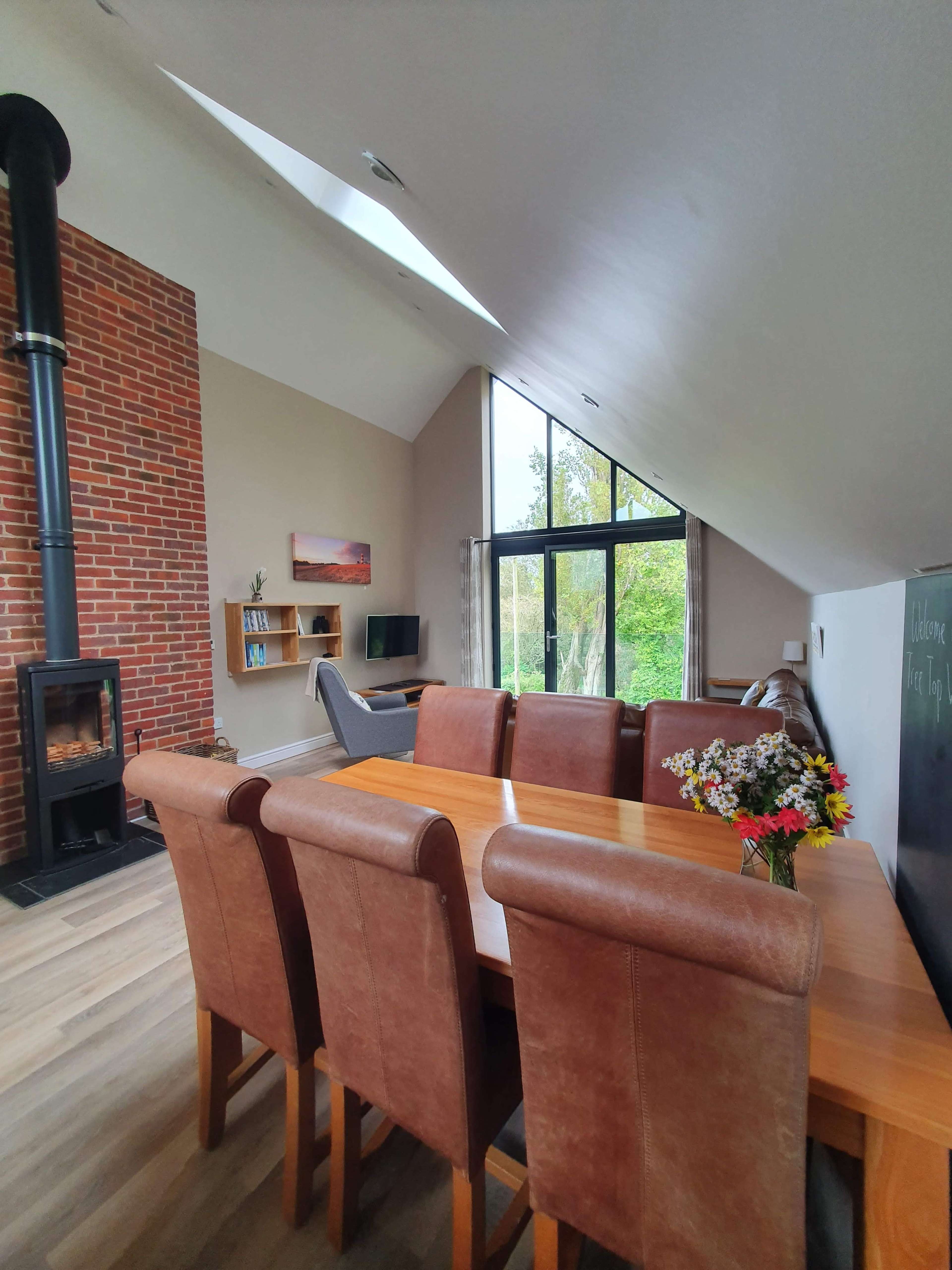 The image shows a modern living space featuring a dining area with a wooden table and brown leather chairs, a cozy fireplace on a brick wall, and large windows overlooking greenery.