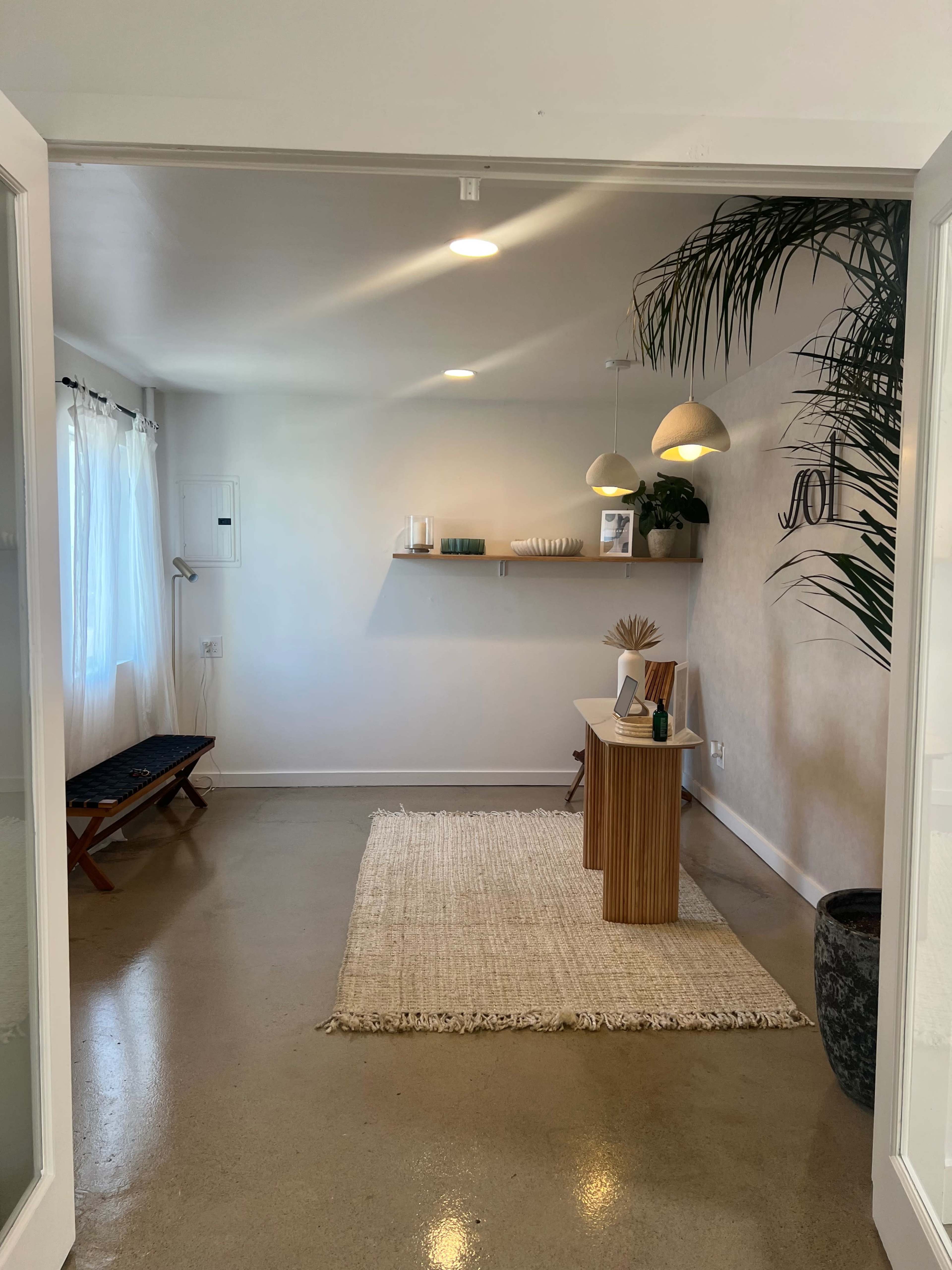 A bright, minimalist room with a wooden table, two pendant lights, a rug on the floor, and a shelf displaying decorative items.