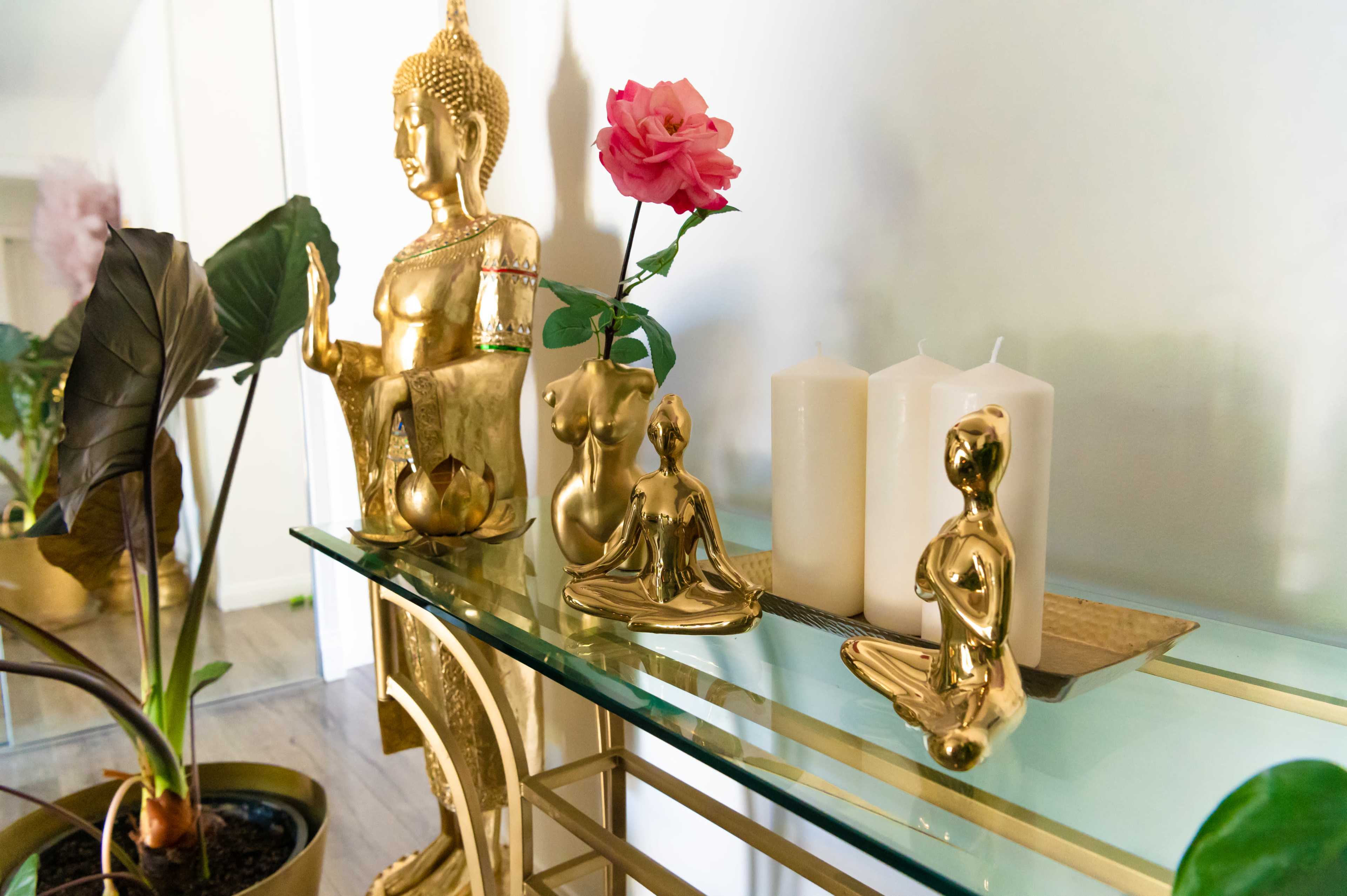 A gold Buddha statue, small golden figurines, a pink rose in a vase, and three white candles displayed on a glass shelf.