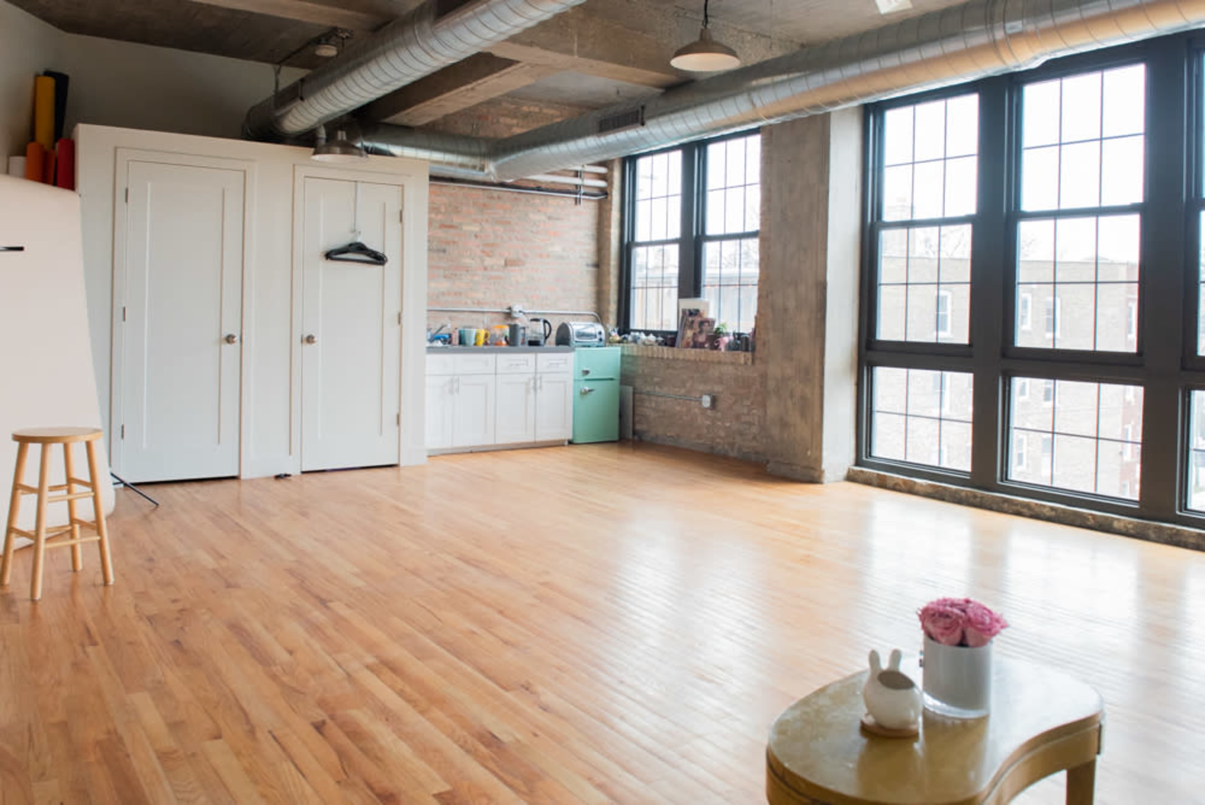 A spacious, modern studio with wooden floors, large windows, and a kitchenette against a brick wall.