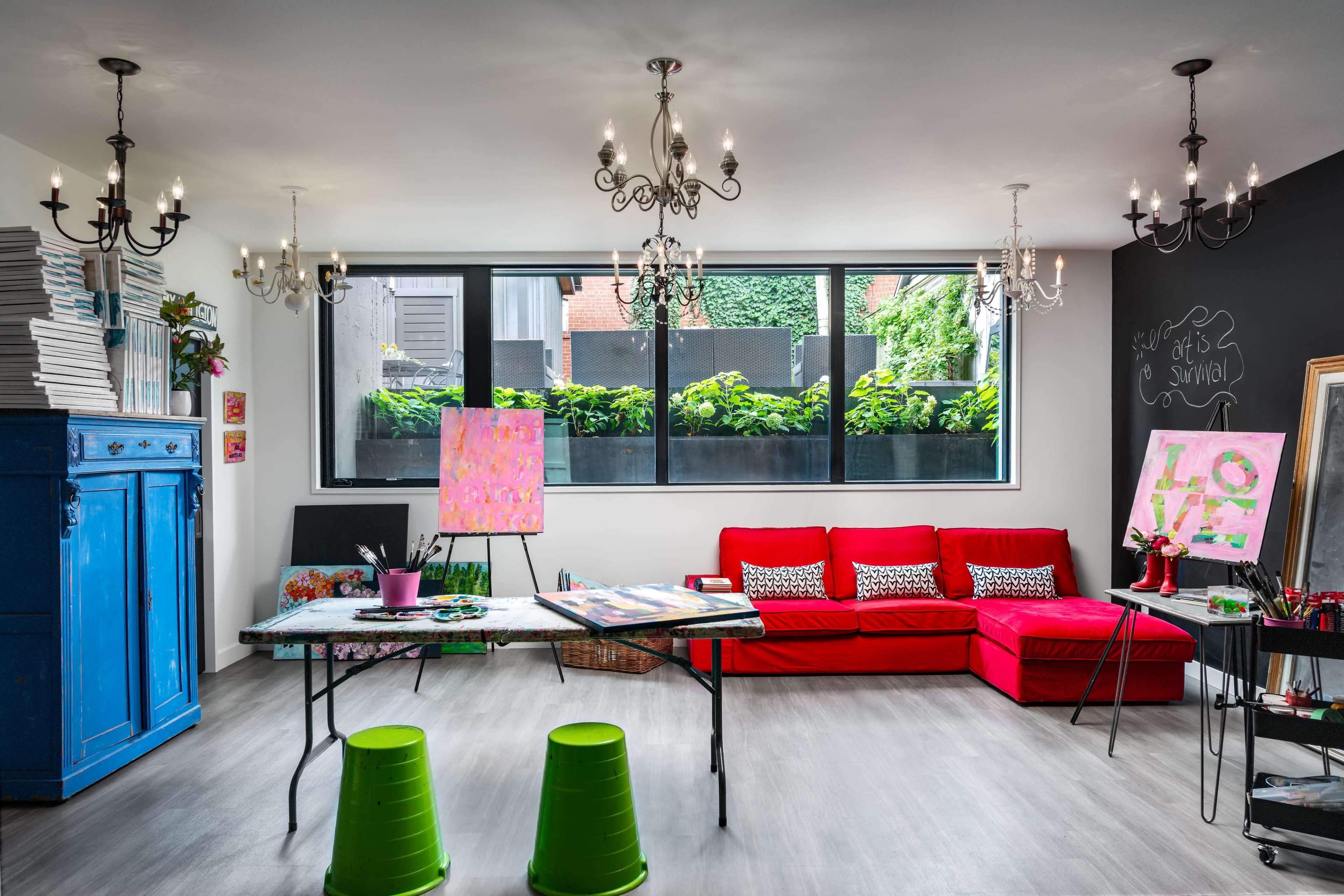A bright and stylish art studio featuring a red couch, a large window with greenery outside, and a central table surrounded by green stools.