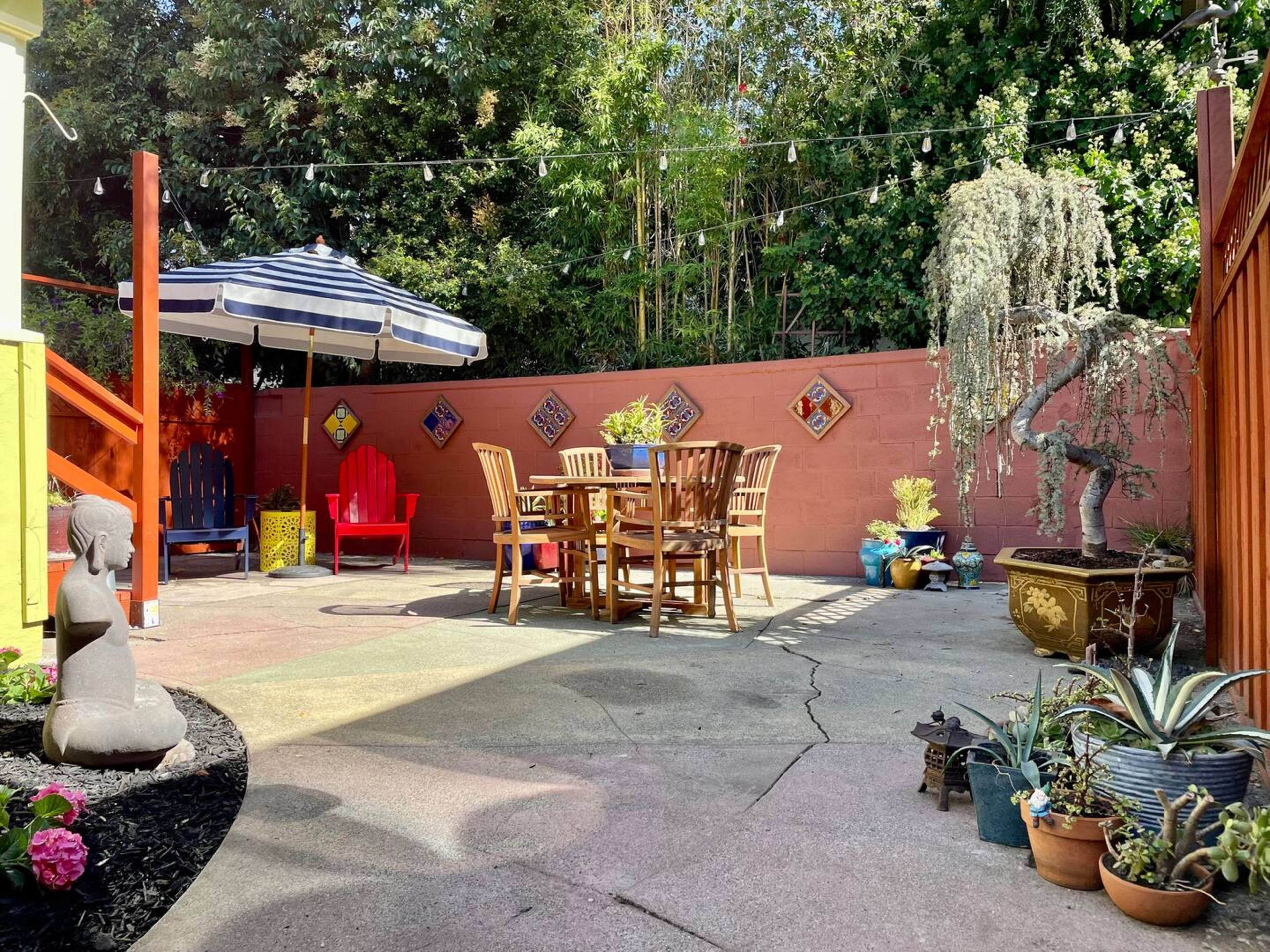 A patio area features a wooden dining set beneath an umbrella, surrounded by decorative plants and artwork on a brick wall.