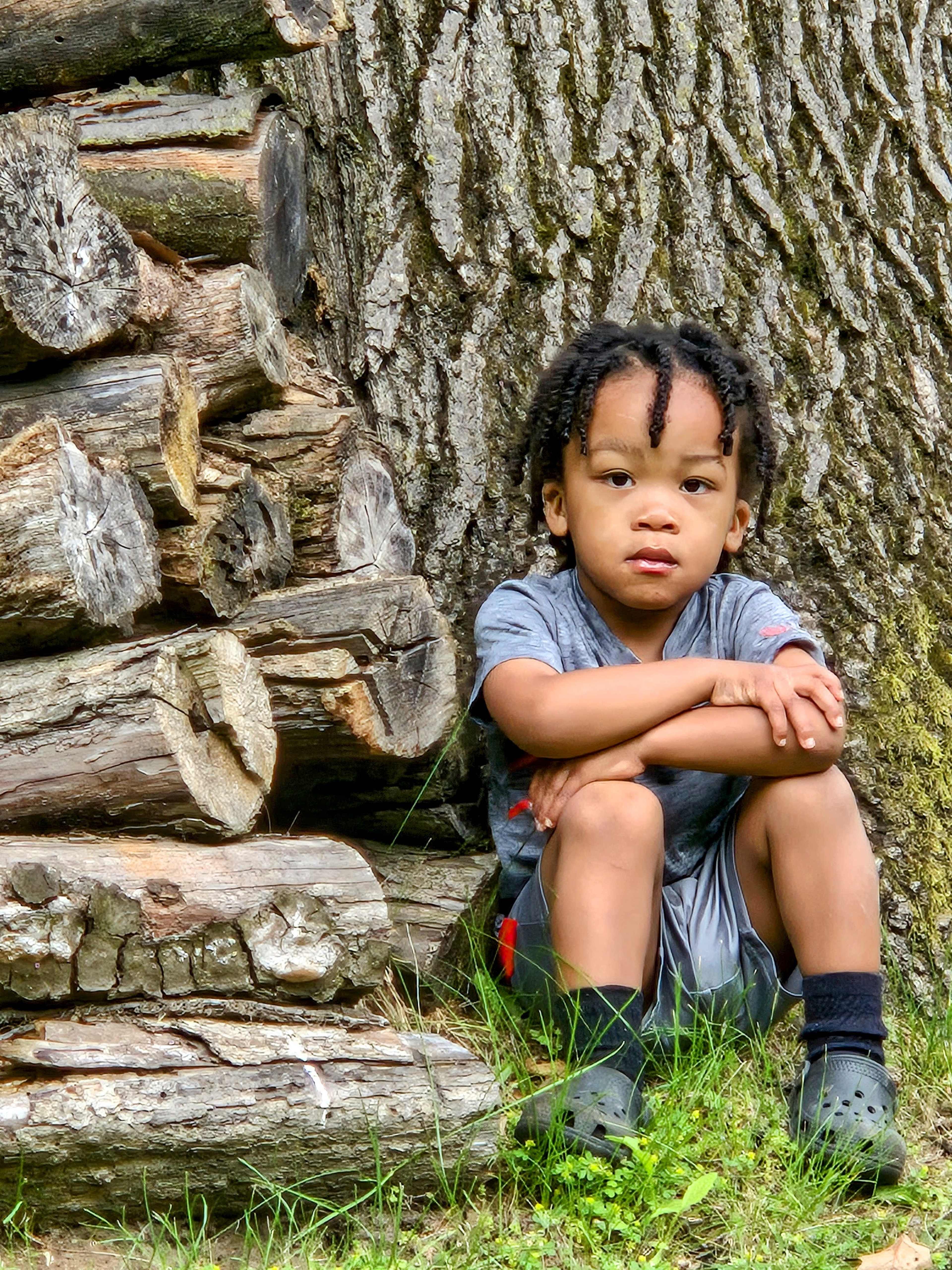 A young child sits on the grass next to a stack of logs, leaning against a tree trunk.