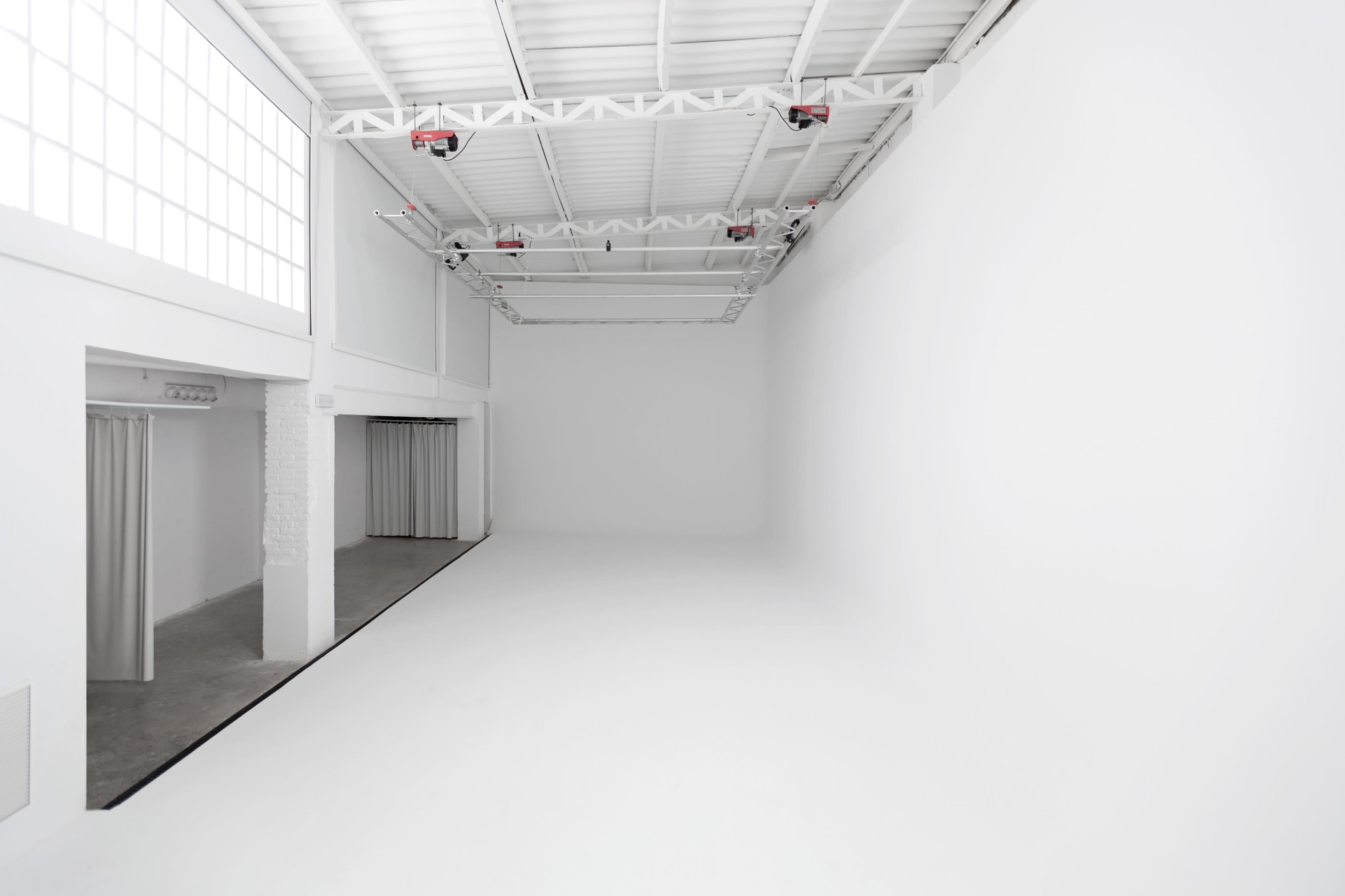 The image depicts a spacious, minimalist studio with white walls, a high ceiling, and an expansive, unobstructed floor area.