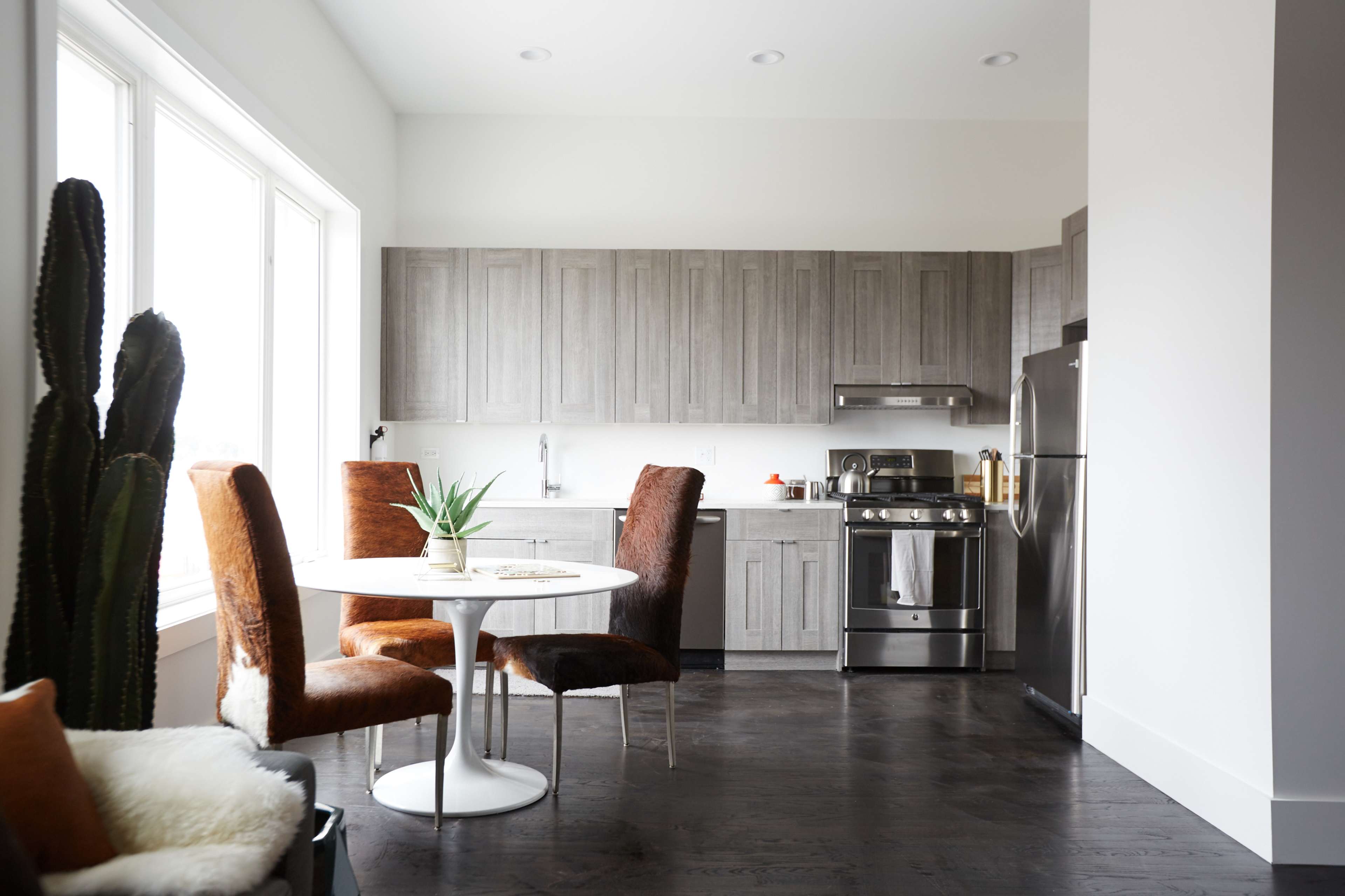 A modern kitchen features gray cabinetry, stainless steel appliances, a round white table with two chairs, and a window allowing natural light.