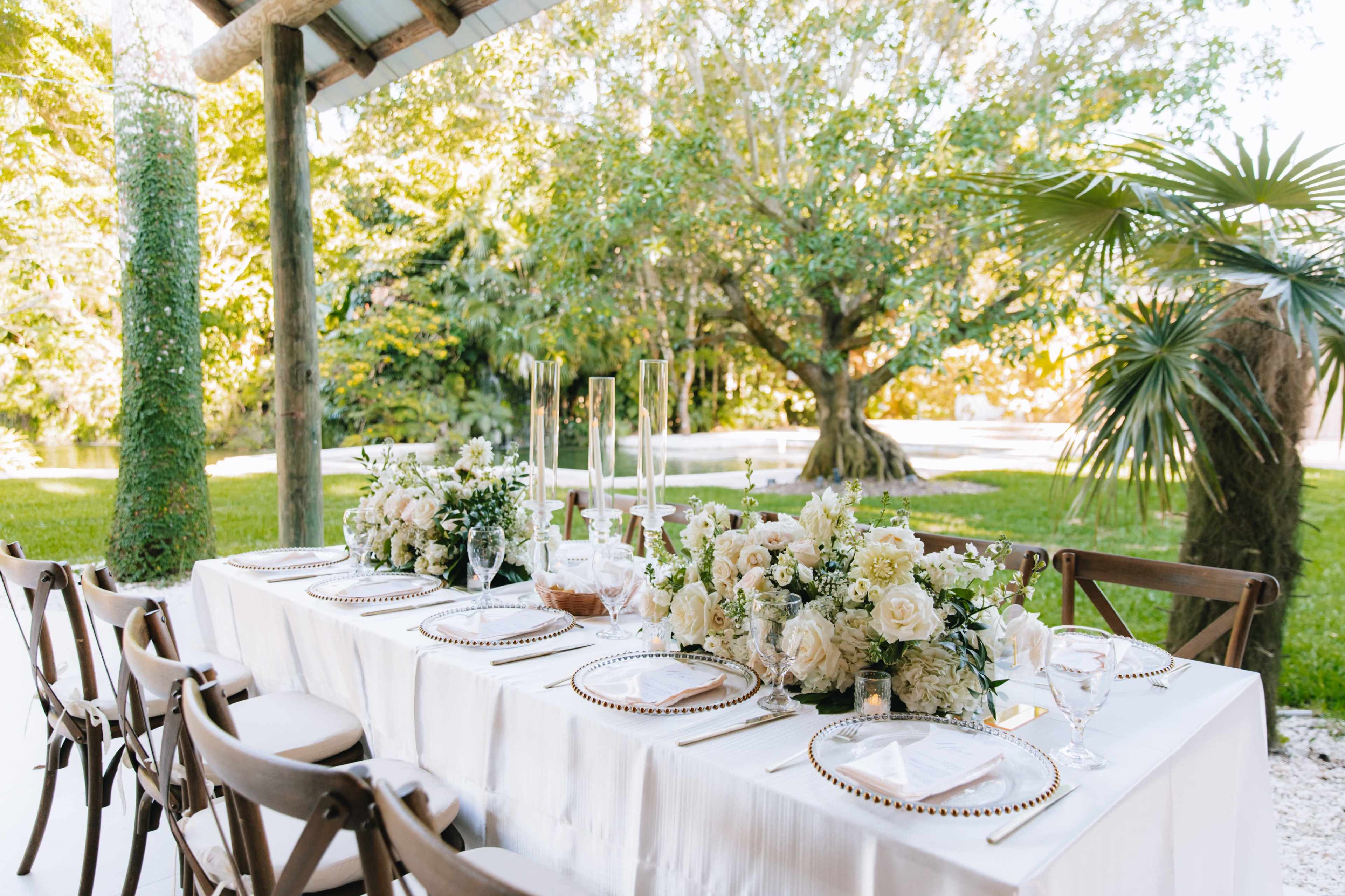 A long table is elegantly set with white floral arrangements and glassware under a shaded outdoor setting surrounded by greenery.
