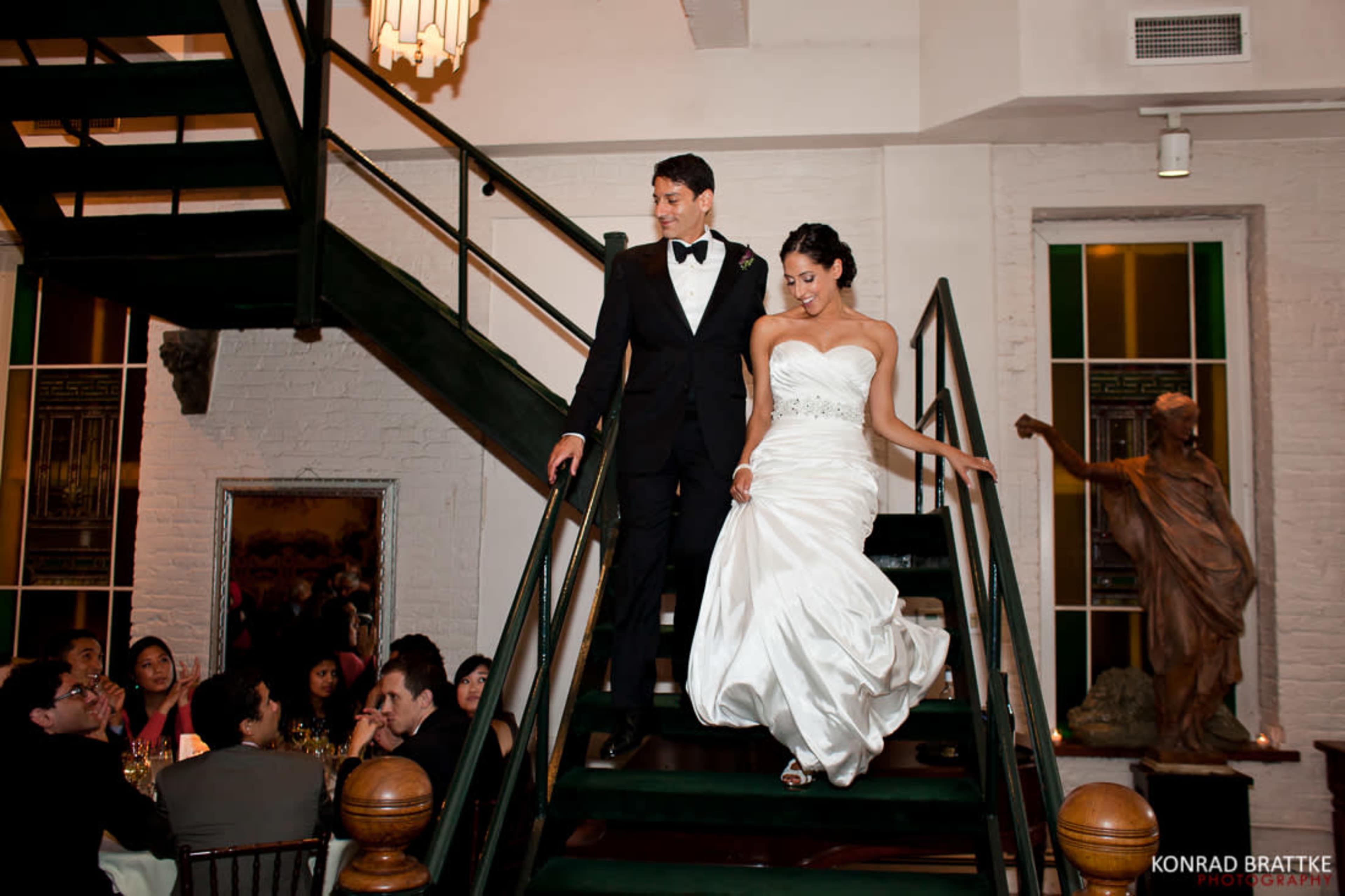 A bride and groom descend a staircase in a banquet hall while guests sit at tables below.