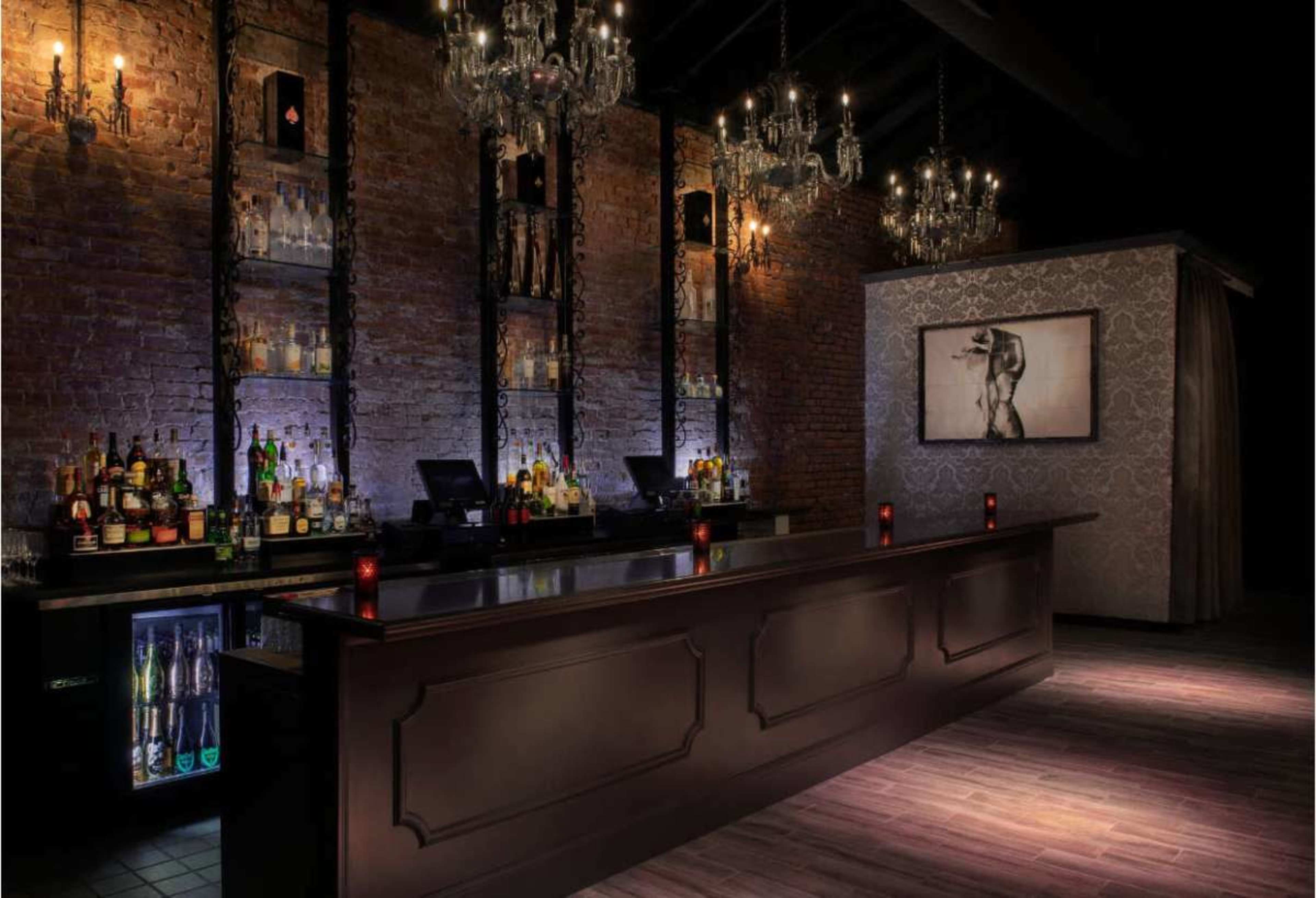 The image shows a dimly lit bar with a dark wooden counter, brick walls, shelves filled with liquor bottles, and elegant chandeliers overhead.