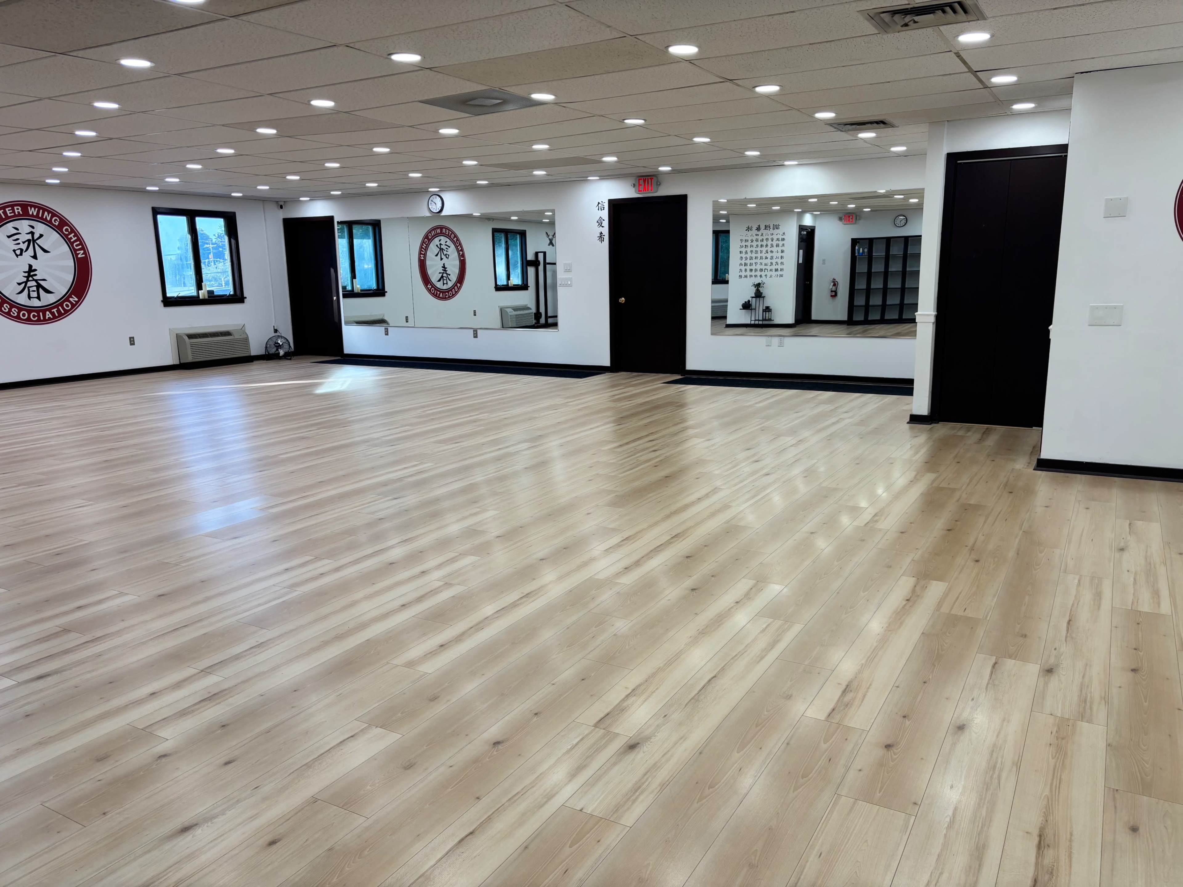 The image shows an empty room with wooden flooring, mirrors on the walls, and wall decorations related to martial arts.