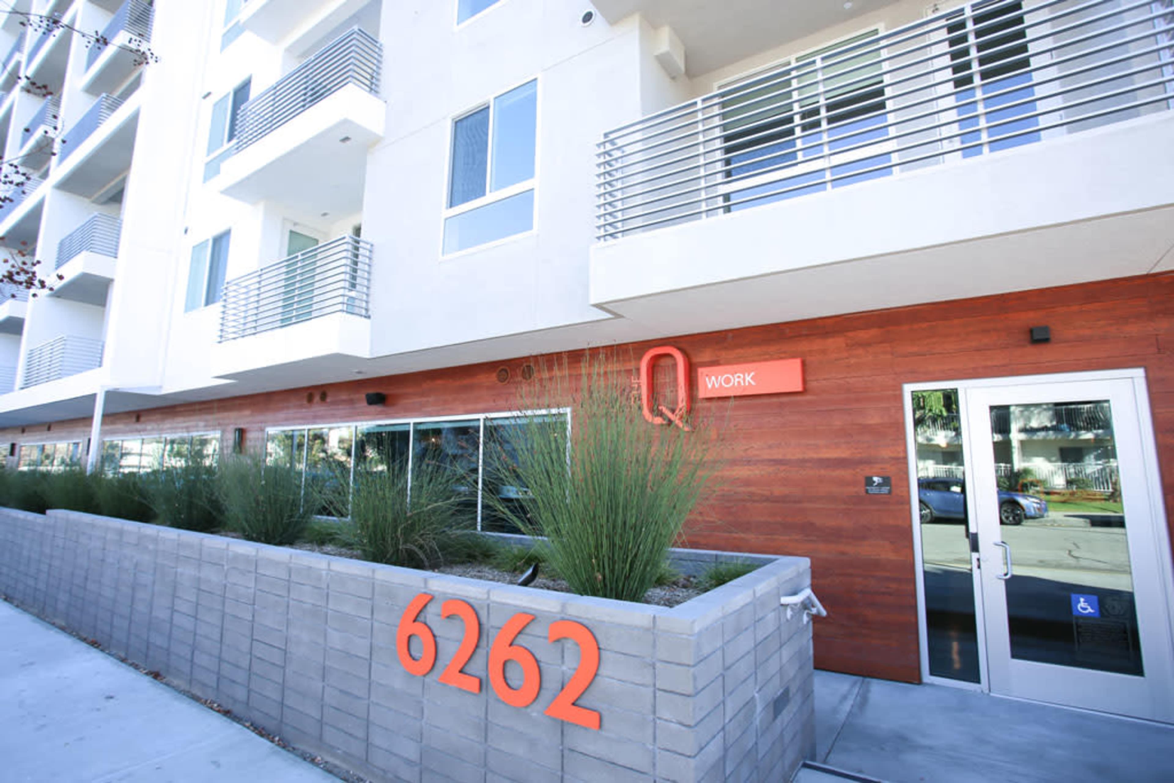 A modern building facade with a sign reading "Q WORK" and a planter containing tall green grass in front of it.