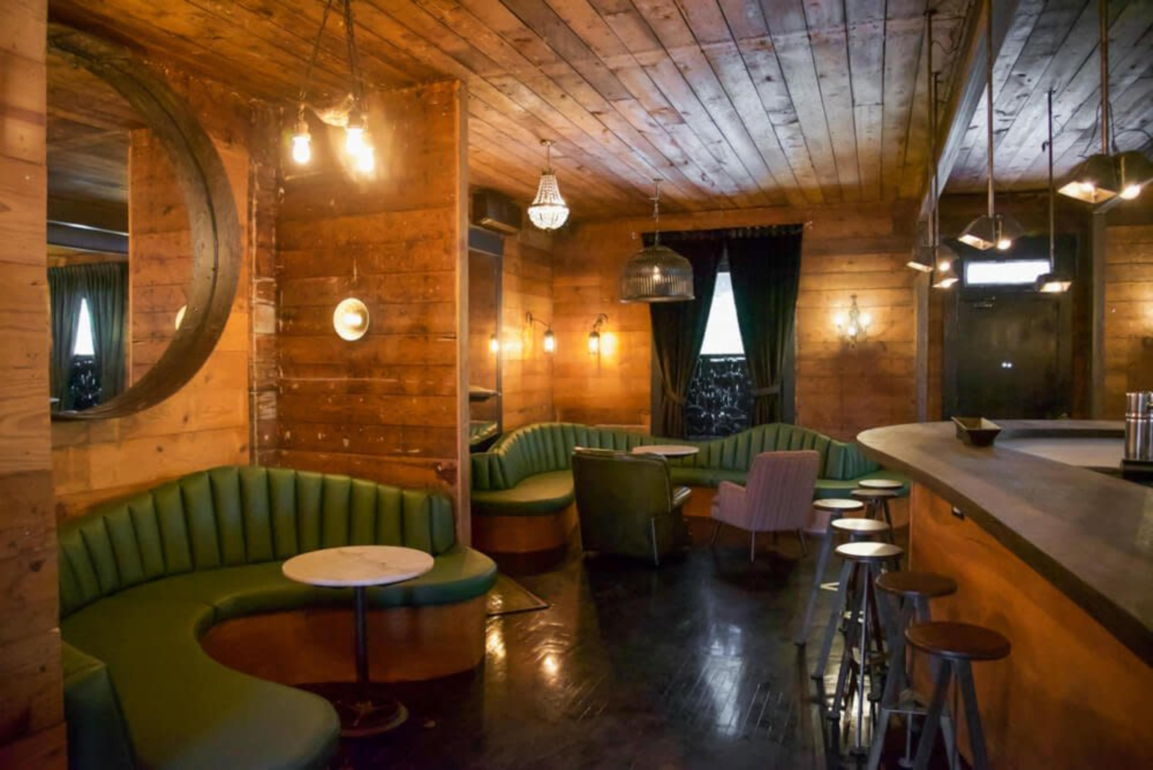 The interior of a cozy bar features wooden walls, green curved seating, and various light fixtures illuminating the space.