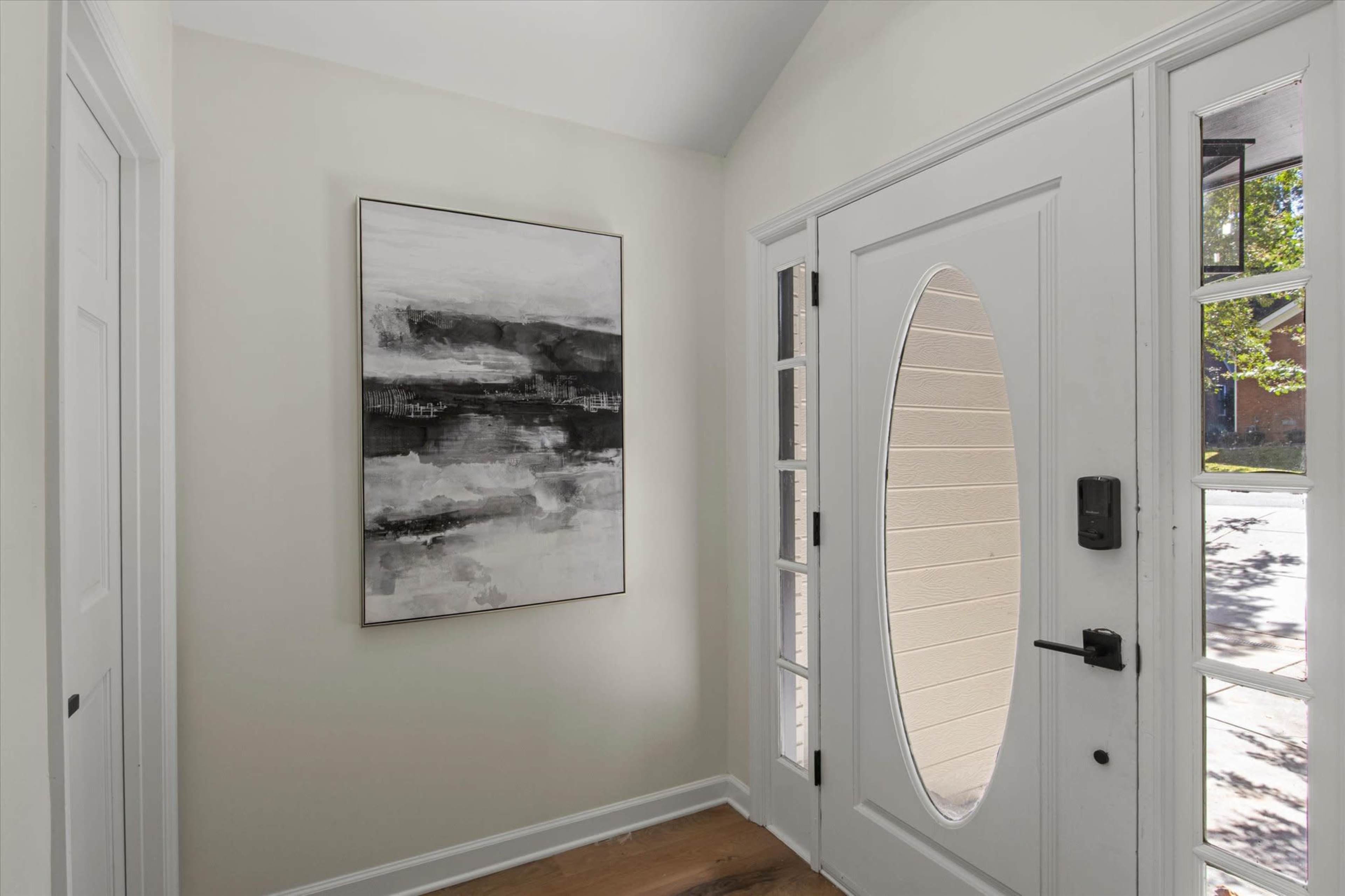 The image shows an entrance hall featuring a white door with an oval window, a black-and-white abstract painting on the wall, and a glimpse of natural light through a nearby window.