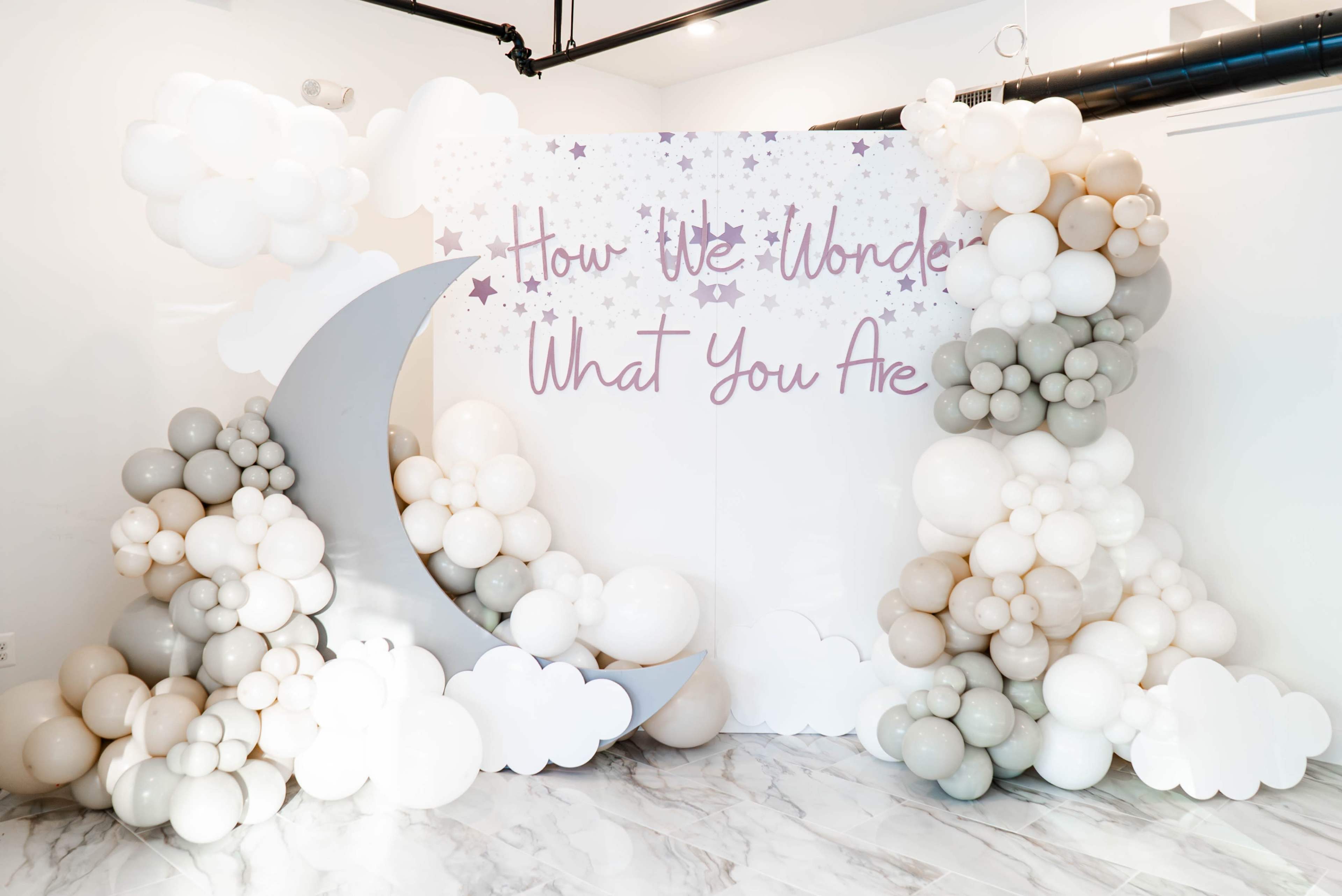 A decorative backdrop with a crescent moon and clusters of balloons in soft colors, adorned with the phrase "How We Wonder What You Are."