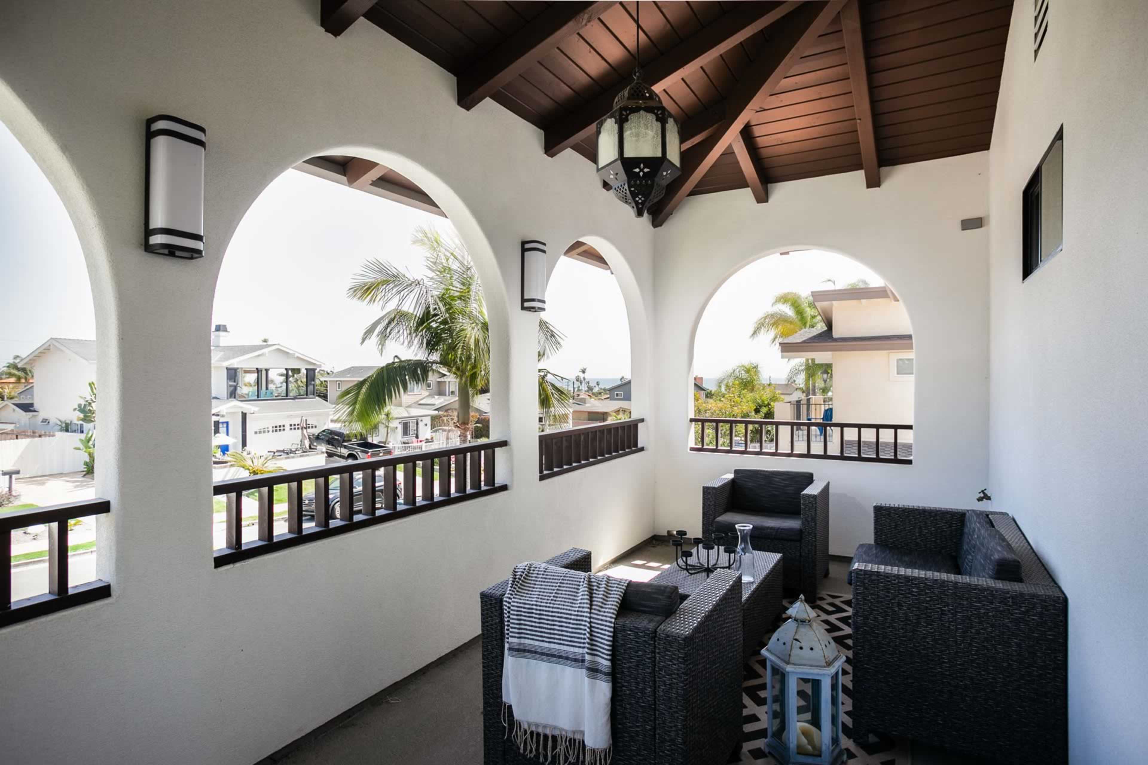 A covered balcony with arched openings, featuring wicker furniture, a lantern, and decorative tiles, overlooking a residential area.