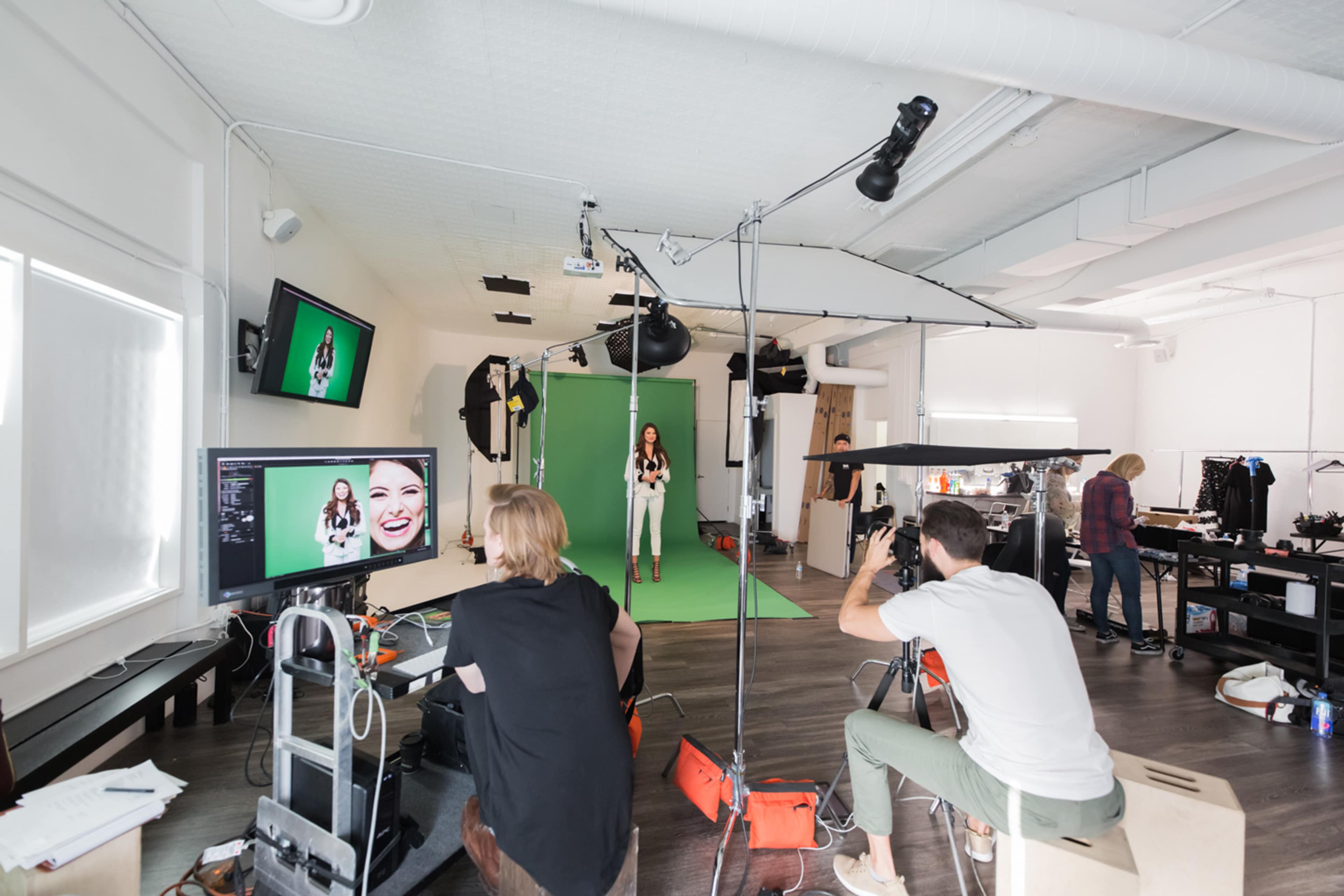 A professional photo shoot is taking place in a studio with a model posing in front of a green screen while photographers and crew work on equipment and monitors.