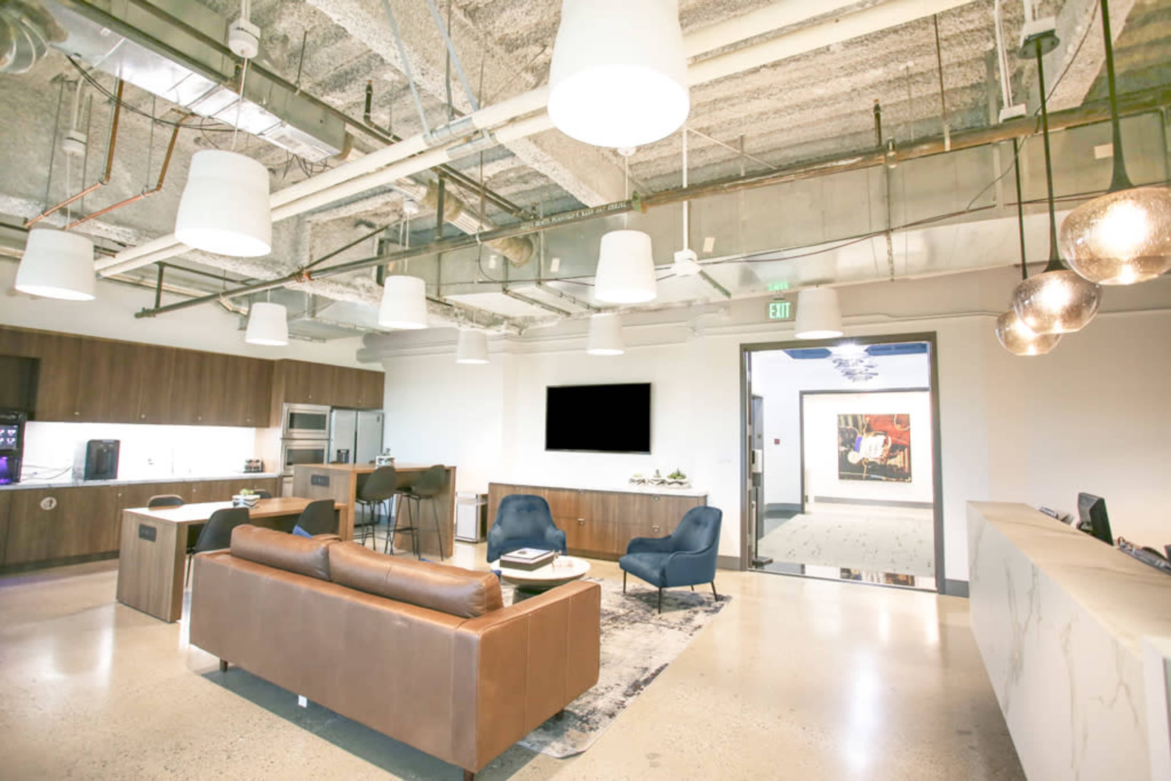 A modern office lounge area with a brown leather sofa, two blue chairs, a kitchenette, and overhead lighting fixtures.