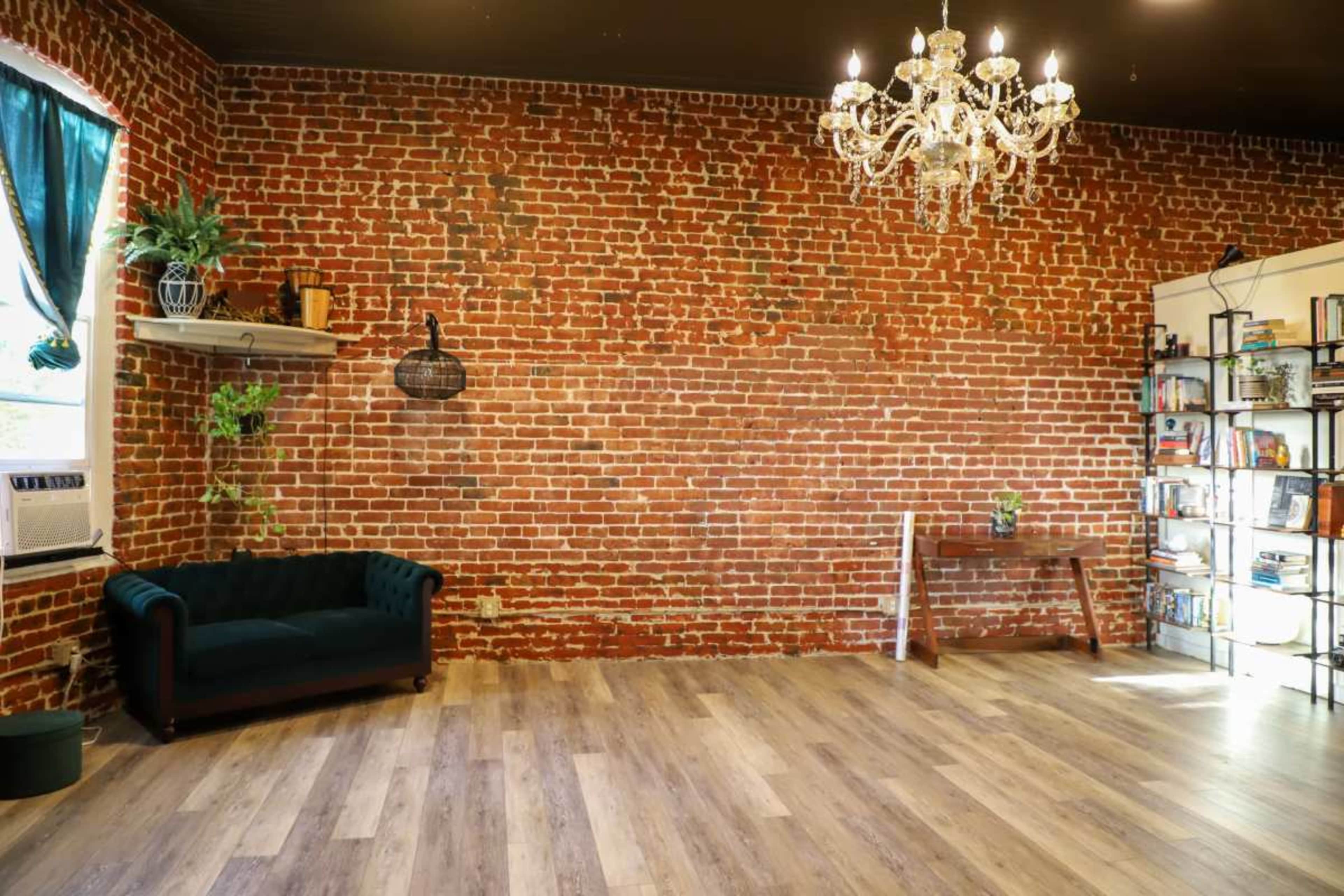 The image shows a room with a brick wall, a green couch, a wooden table, a bookshelf filled with books, and a chandelier.