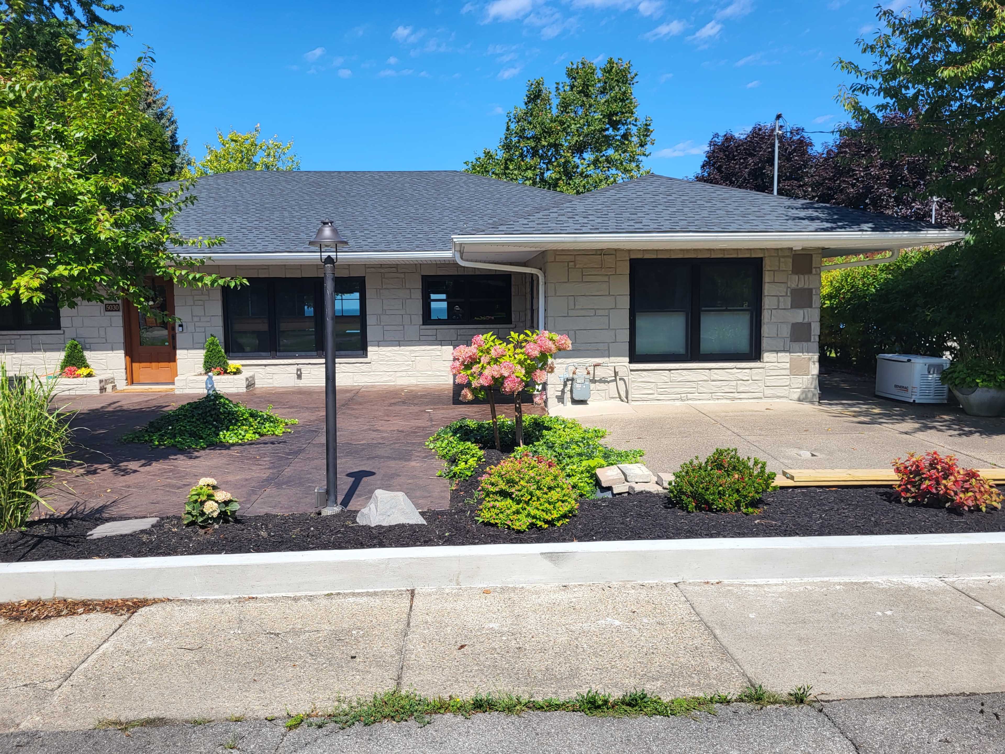 A stone-faced house with a landscaped front yard featuring flowers, shrubs, and a paved walkway.
