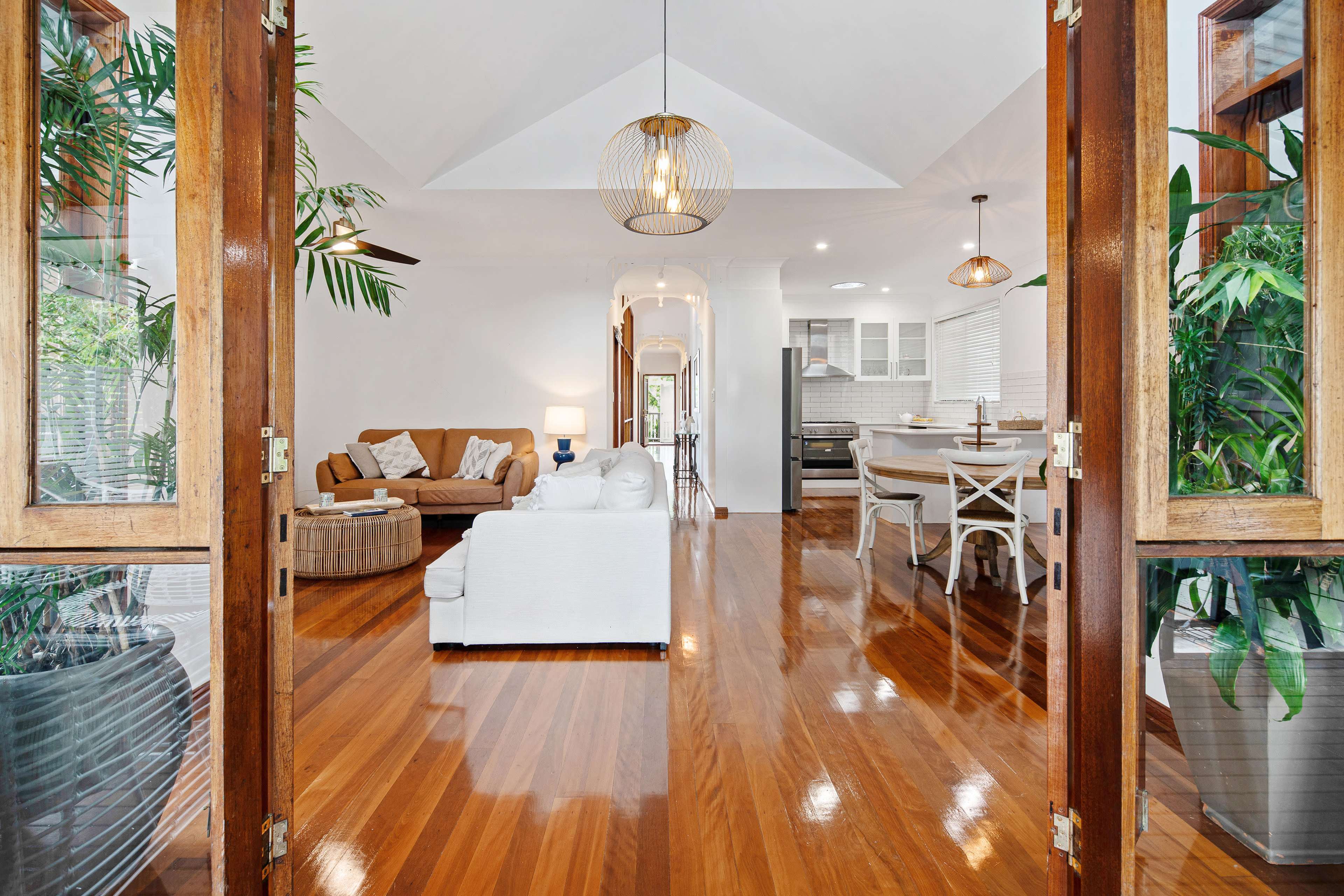 A living room features wooden flooring, a white sofa, brown chairs, and large plants, with light streaming through open double doors.