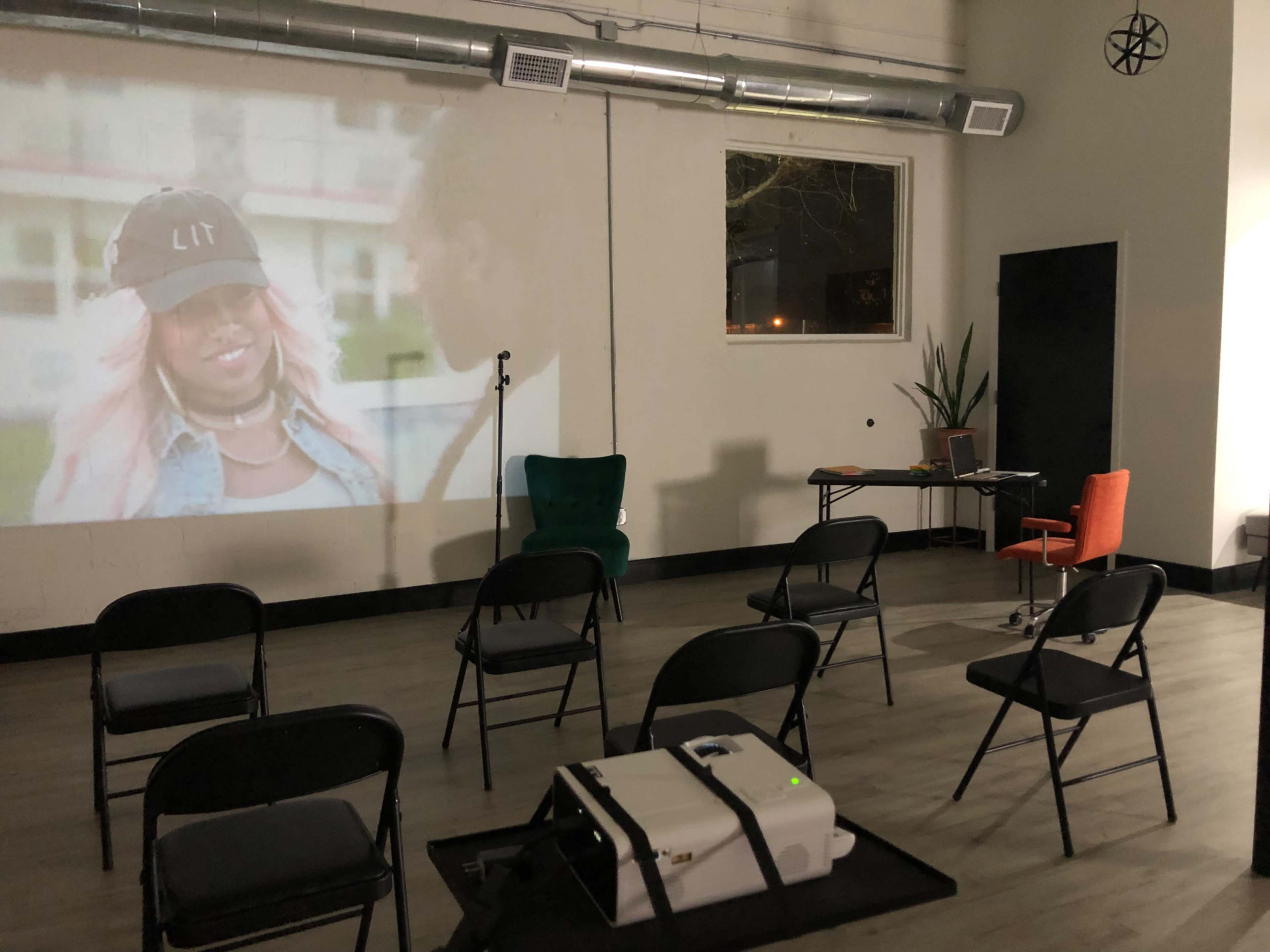 A projection screen displays a video while chairs are arranged in a semi-circle around a table and a microphone stand in a sparsely decorated room.